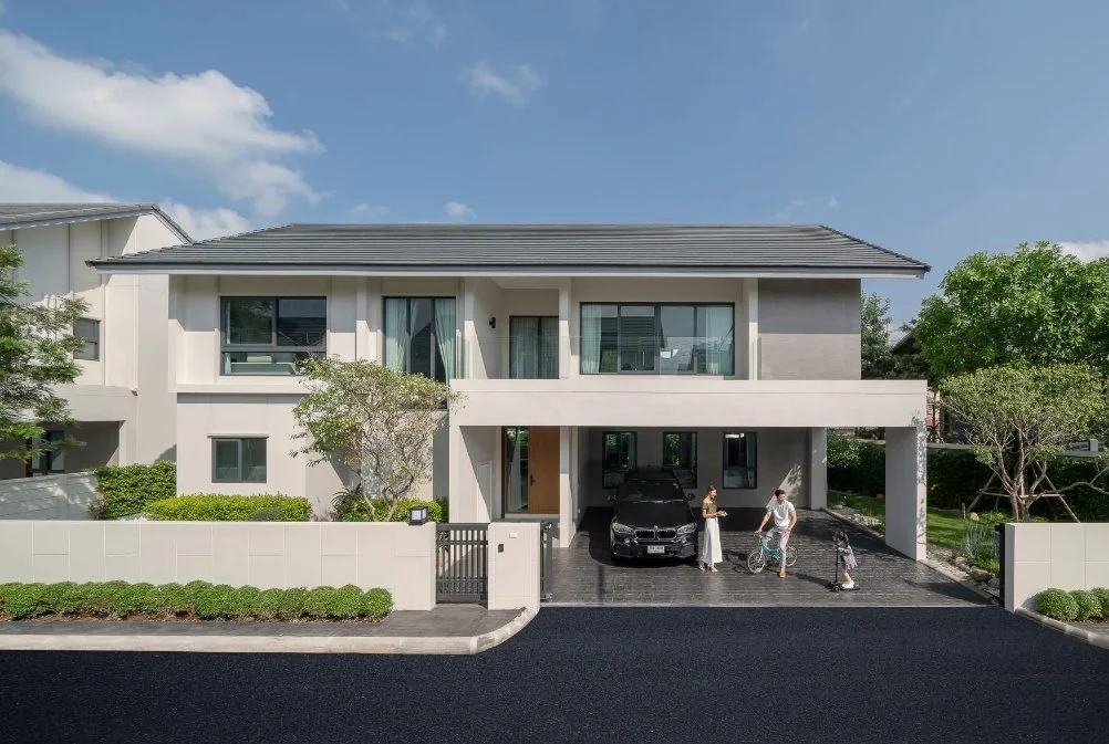 Modern two-story house with a gray tiled roof, white exterior walls, large windows, and a front porch. There are two people standing near the driveway, one with a bicycle and electric scooter, and a black car parked under the porch. Green trees and bushes are around the property, with a blue sky and some clouds overhead.