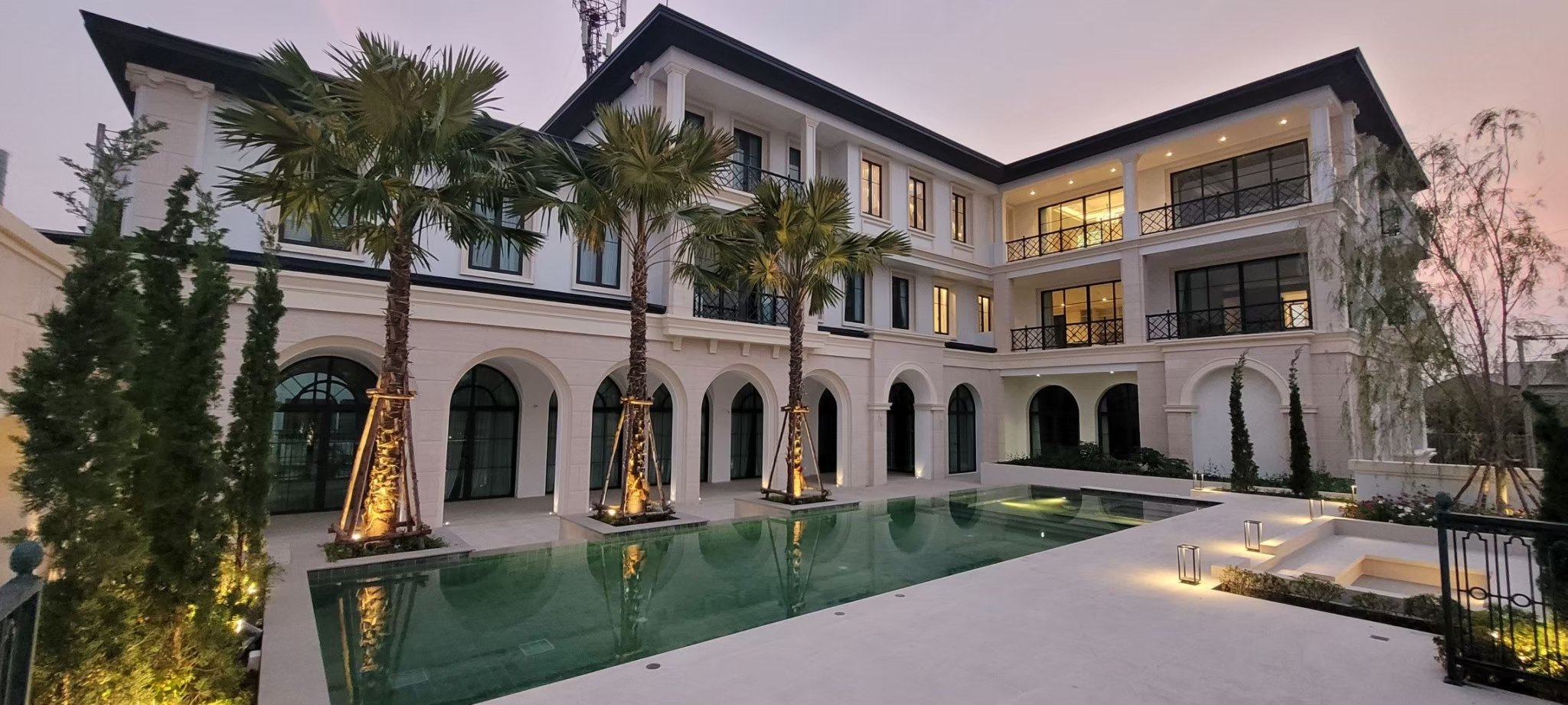 Luxury multi-story house with a swimming pool, palm trees, and outdoor lighting during twilight.