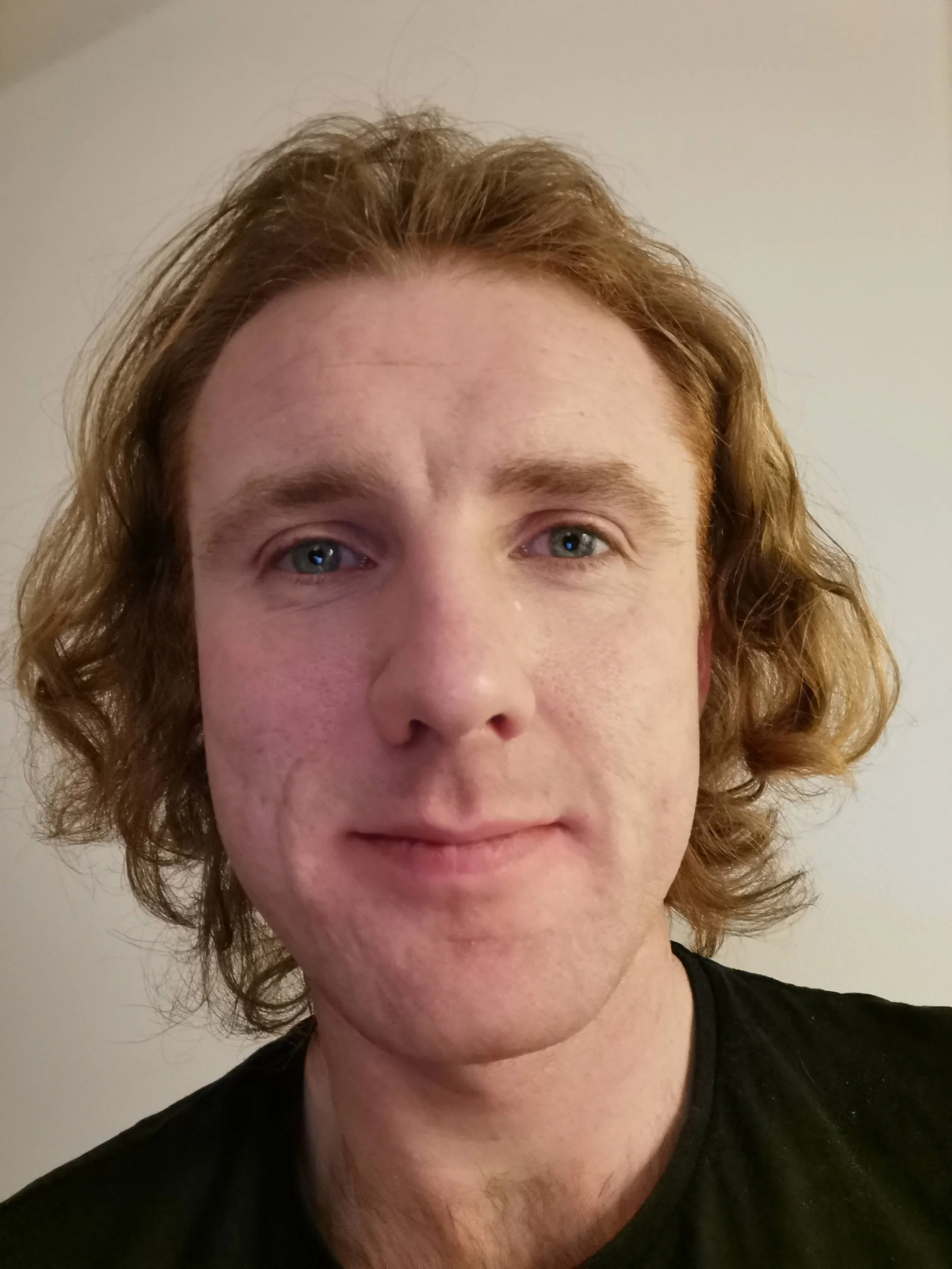 Alba Automations Member: Close-up of a young man with curly red hair and blue eyes, wearing a black shirt, standing against a plain wall.