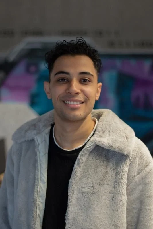 Young man with curly dark hair smiling, wearing a black shirt and light gray hoodie, outdoors next to a car with a colorful graffiti background.