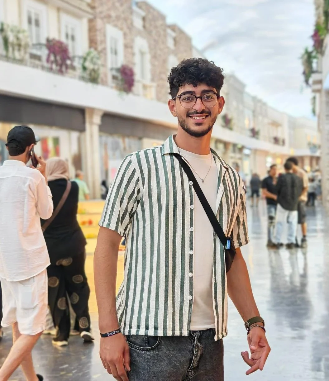 A young man with glasses and a beard smiling, standing on a busy street with shops and other people walking in the background.