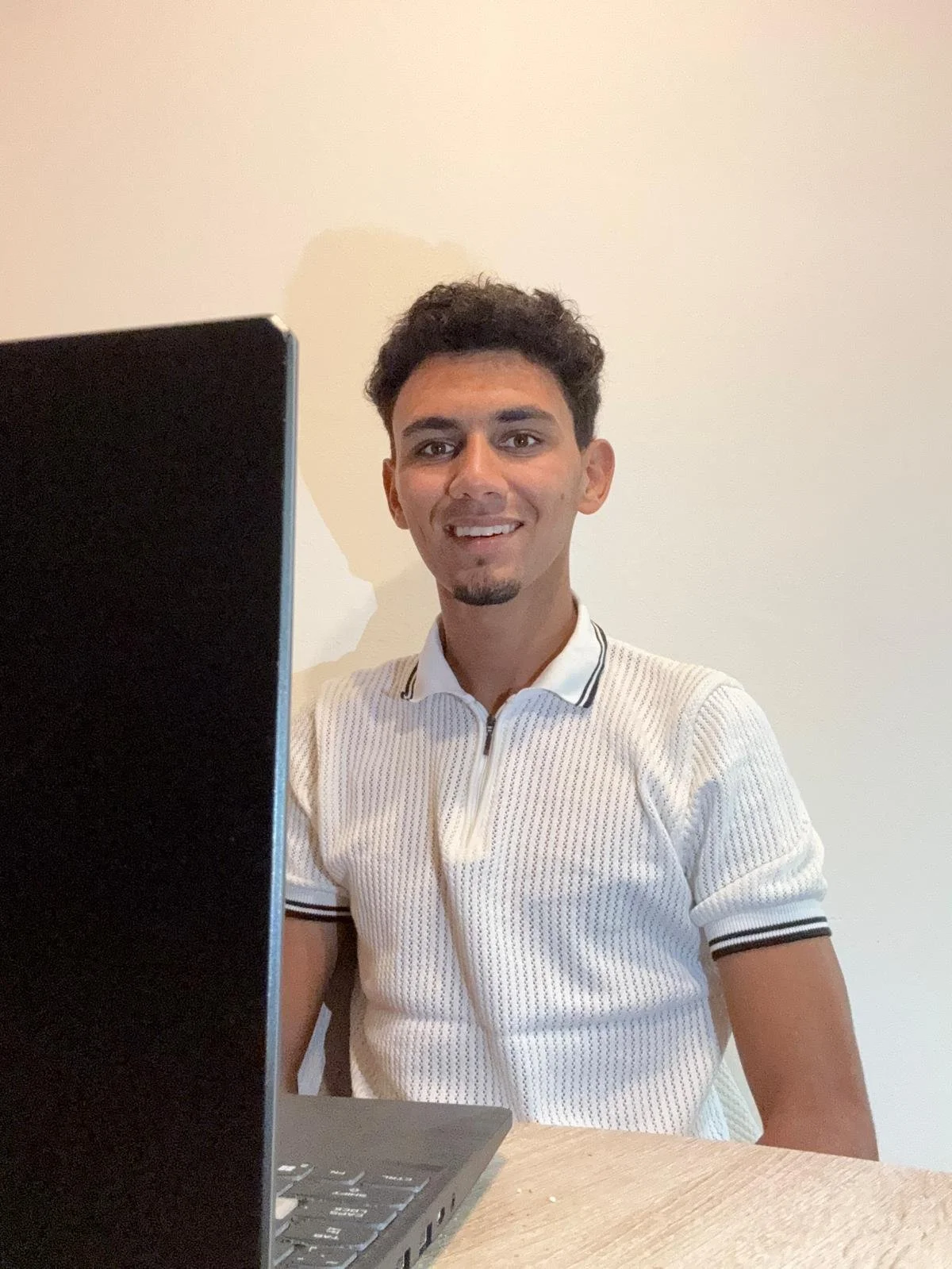 A young man with dark curly hair and a trimmed beard smiling at the camera. He is sitting at a table with a laptop in front of him, wearing a white zip-up polo shirt with black and white striped details.