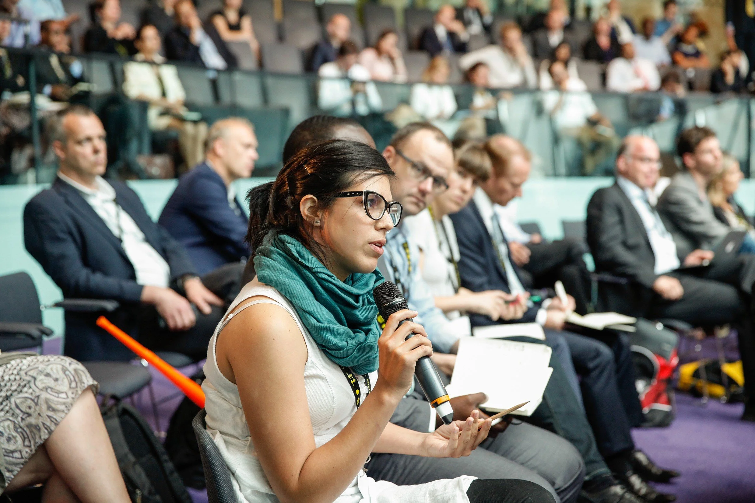 A young woman with dark hair, wearing glasses and a teal scarf, speaking into a microphone during a conference or seminar, surrounded by seated attendees.