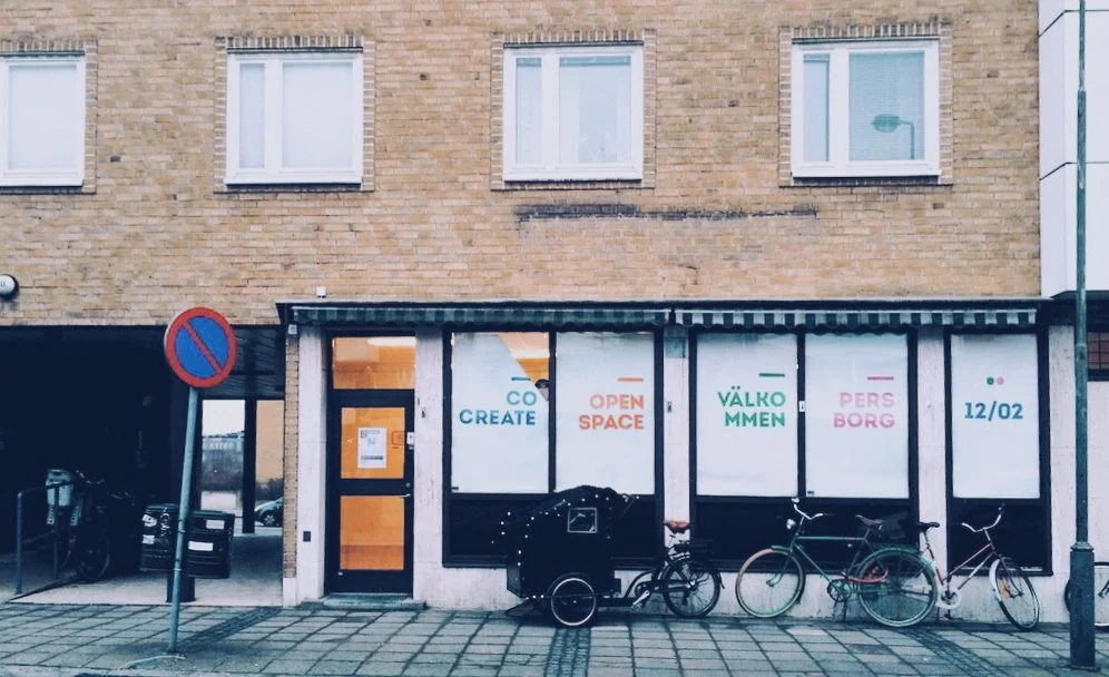 Exterior of a building with large windows, signs for a community space, bicycles parked outside, and a no parking sign.