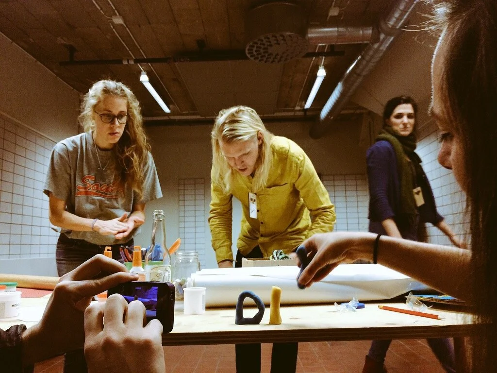 A group of four young women gathered around a table in a room with wooden ceiling and brick walls. The woman on the left with curly blonde hair and glasses is wearing a gray T-shirt, the woman in the center with blonde hair is wearing a yellow shirt