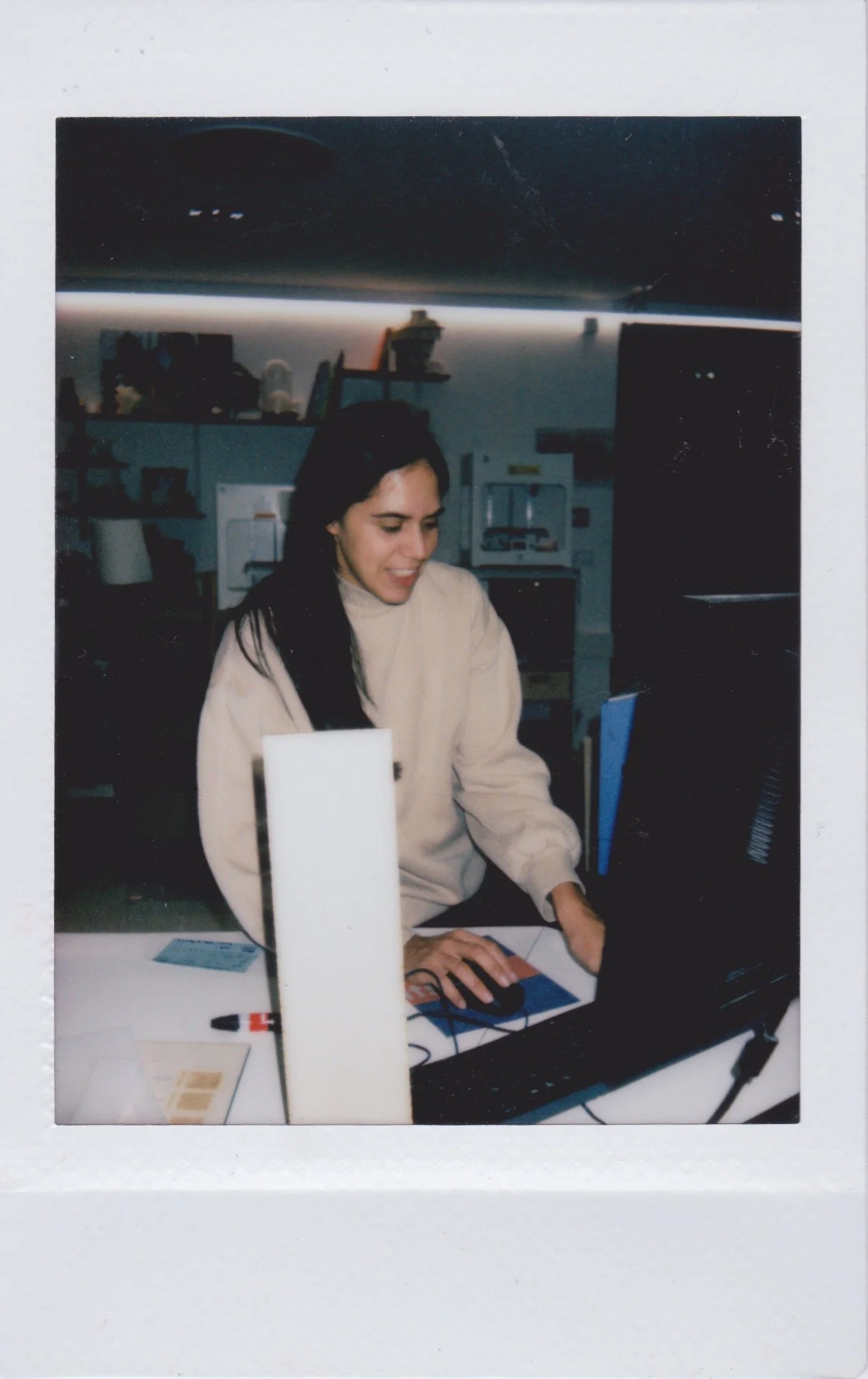 Woman with long dark hair working on a computer in a room with shelves and equipment in the background.
