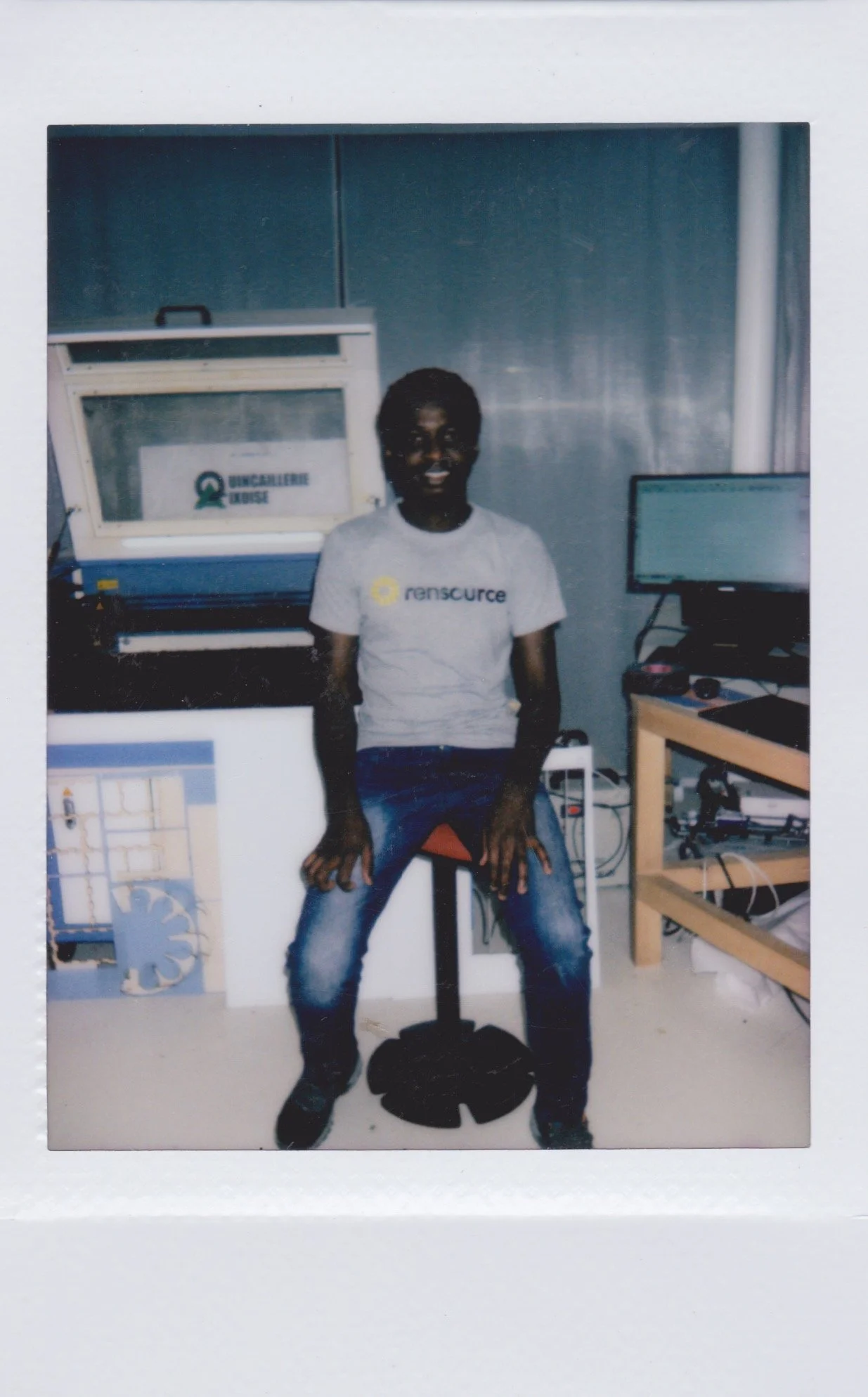 Person sitting on a stool in a tech lab, wearing a t-shirt with 'Rensource' logo, surrounded by computer equipment and a laser cutter in the background.