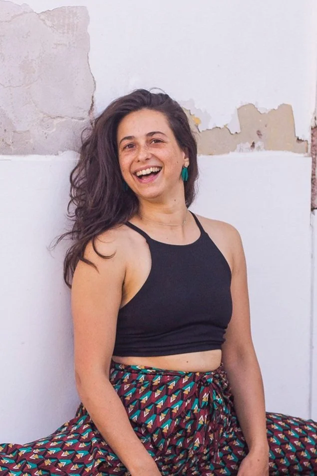 A woman with long, wavy dark hair smiling at the camera, sitting or leaning against a white wall  with peeling paint doing a middle split, wearing a black crop top and multicolored patterned pants.