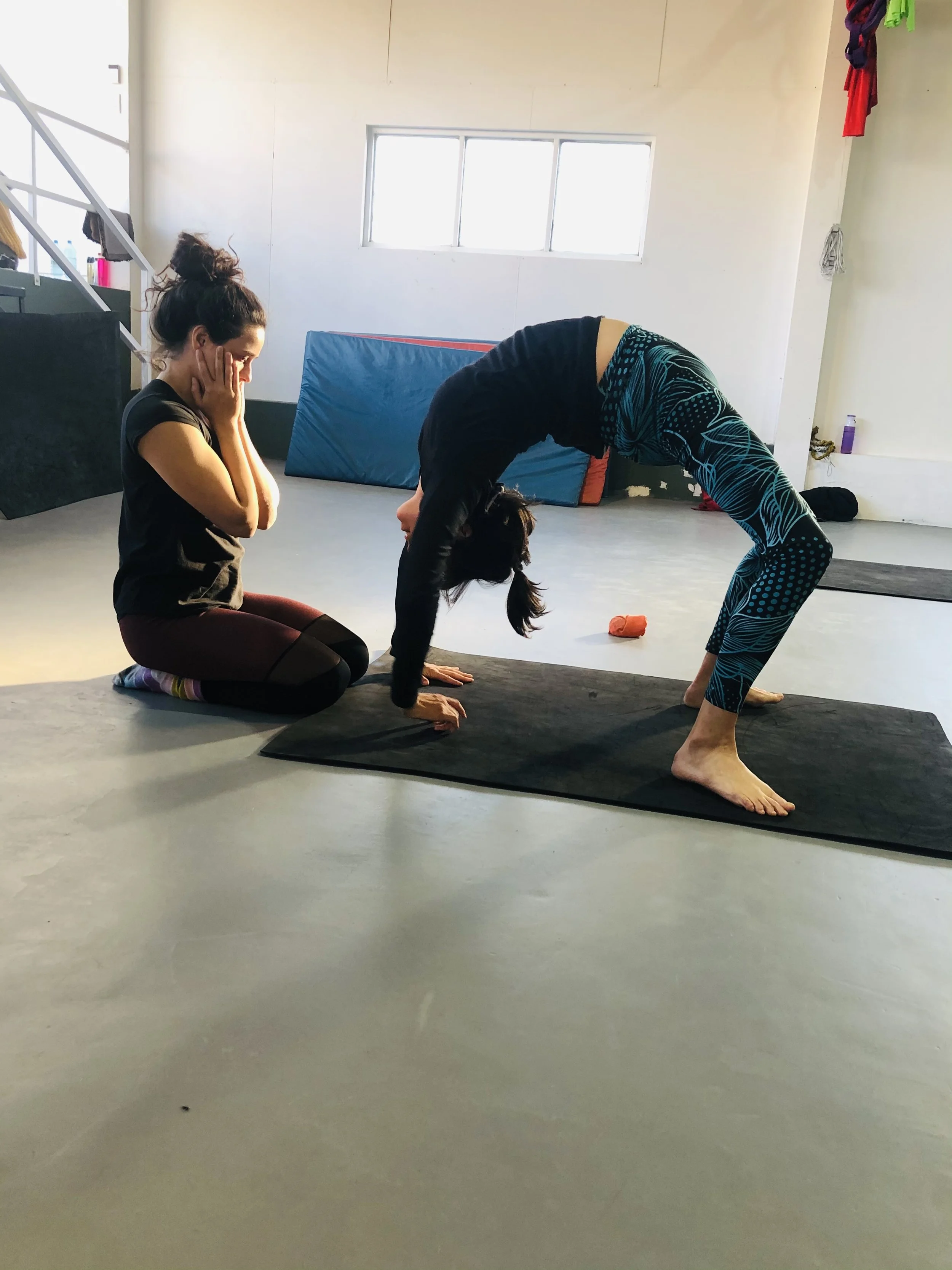 A woman performing a bridge while another woman sits on her knees watching in a gym or studio with mats and fitness equipment. Shoulder stretch and backbend