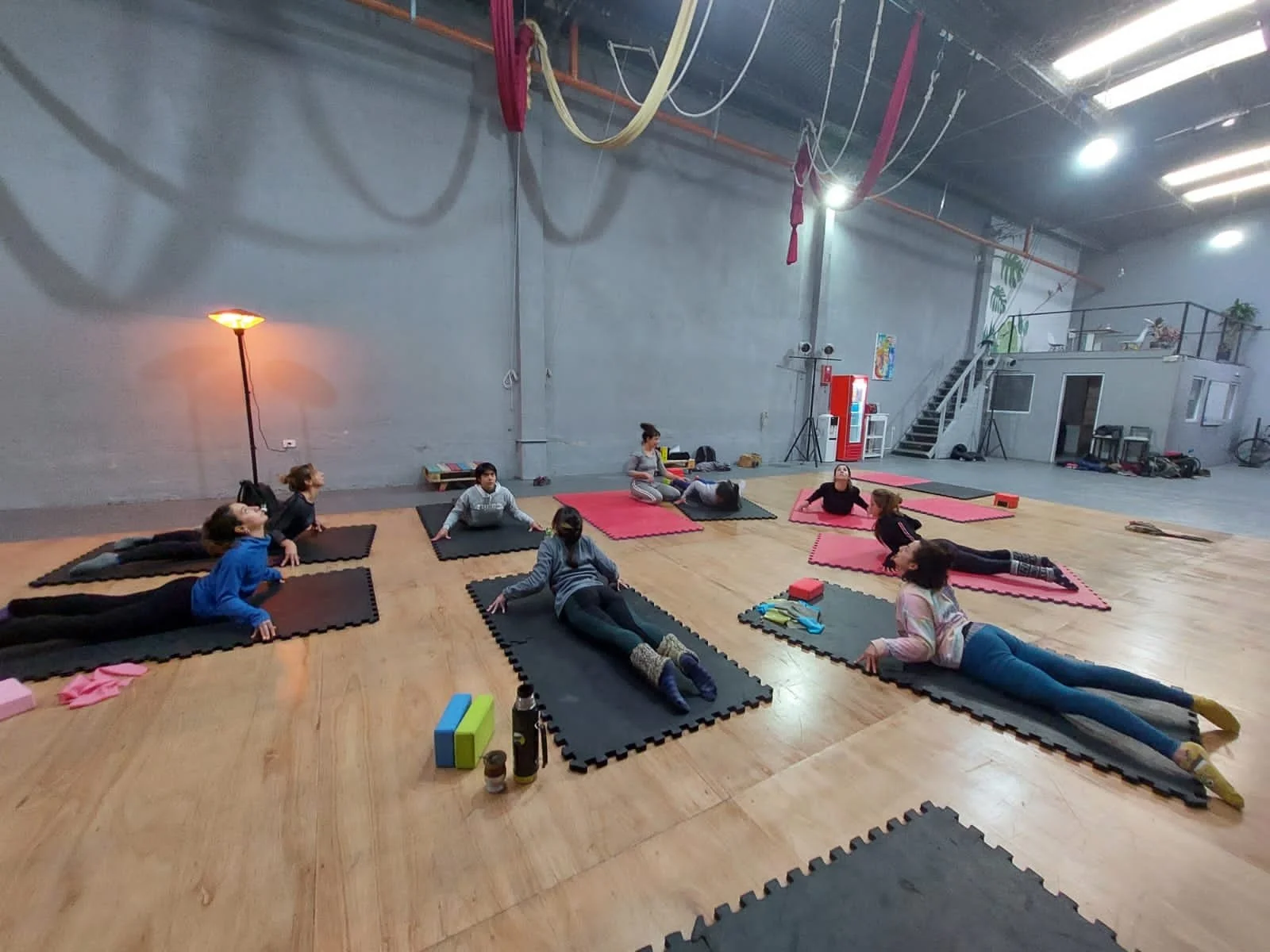 A group of people doing yoga in a spacious studio, lying on mats on a wooden floor, with ropes hanging from the ceiling and a staircase leading to a loft area in the background.