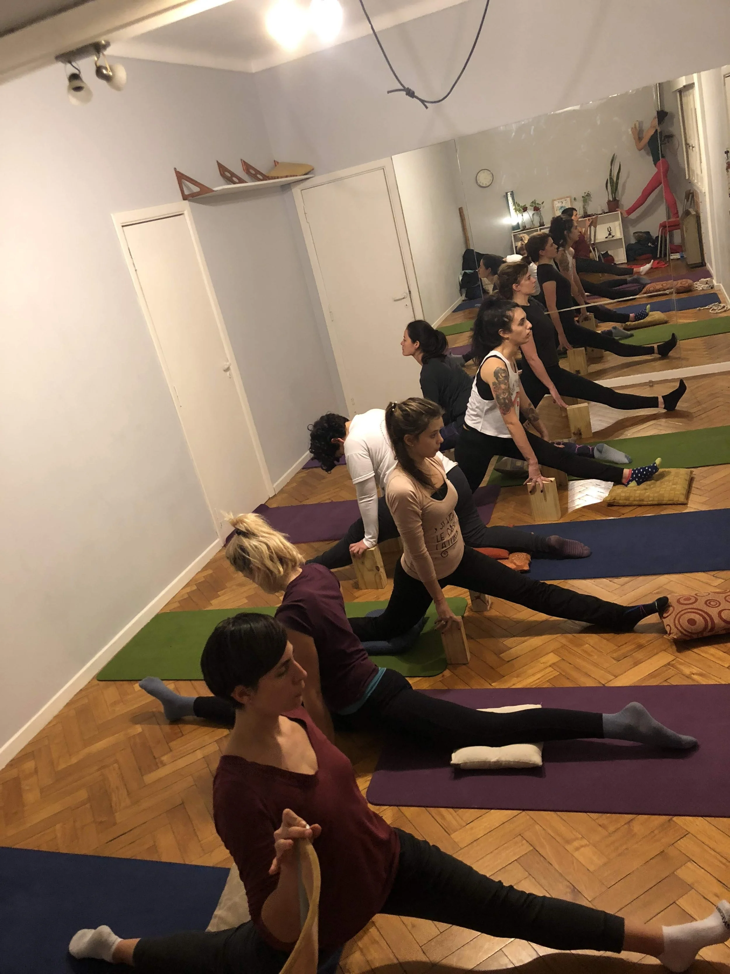 A group of women participating in a yoga class in a studio, practicing stretches with yoga blocks and mats, in front of a mirror.