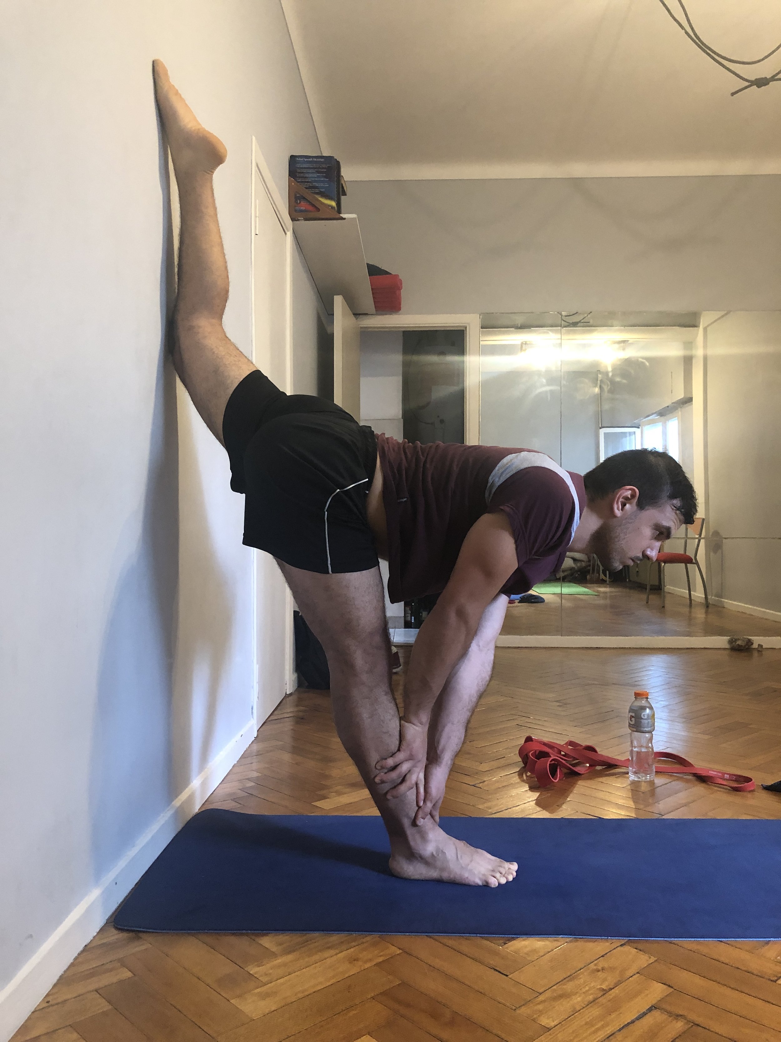 A man practicing standing splits on a wall in a living room, performing a forward bend pose with one leg resting against the wall, wearing black shorts and a maroon t-shirt.
