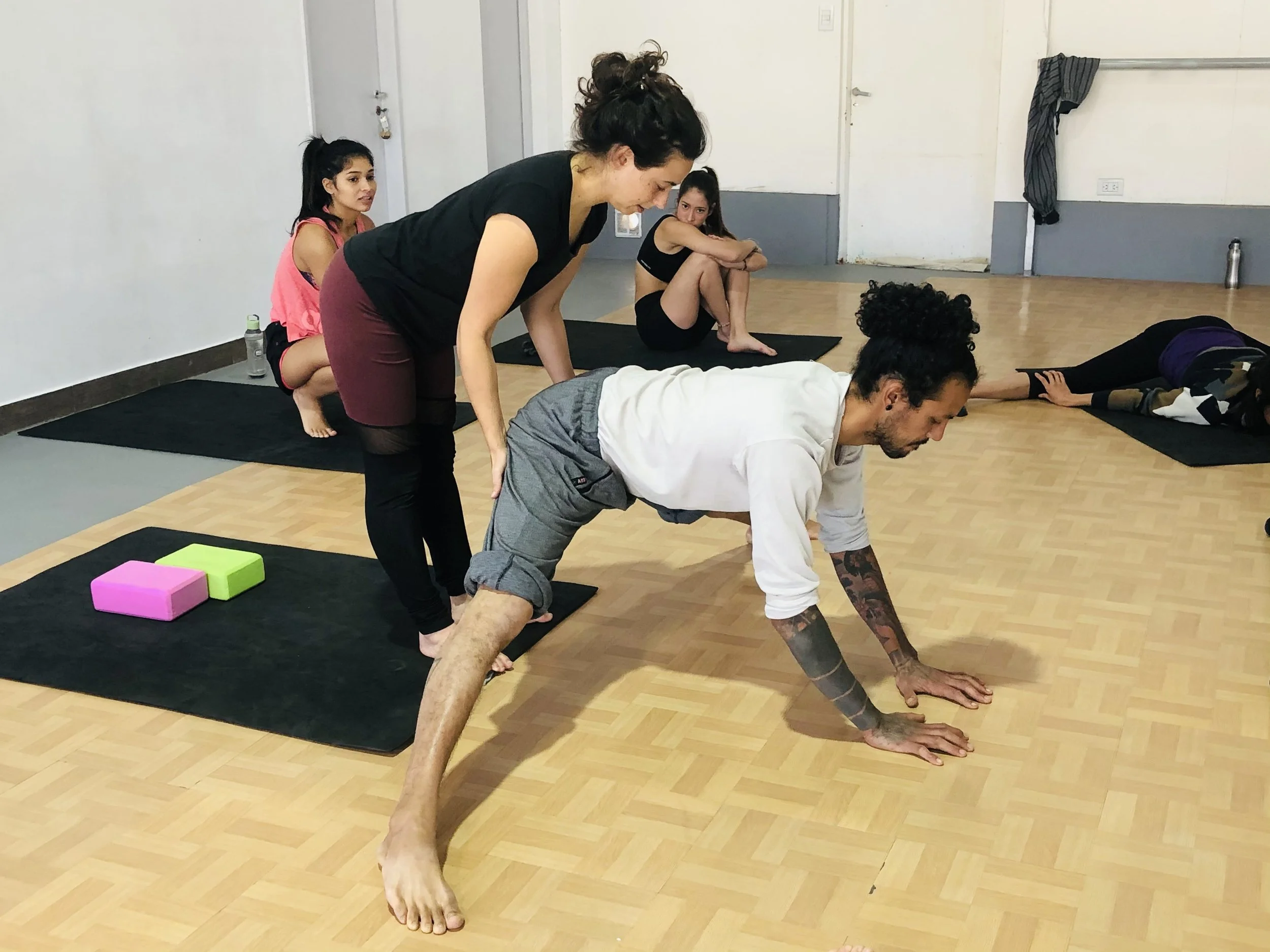 A group of people practicing yoga indoors; a man is practicing midle splits with a trainer supporting their hips, while others are sitting or lying on yoga mats in the background.