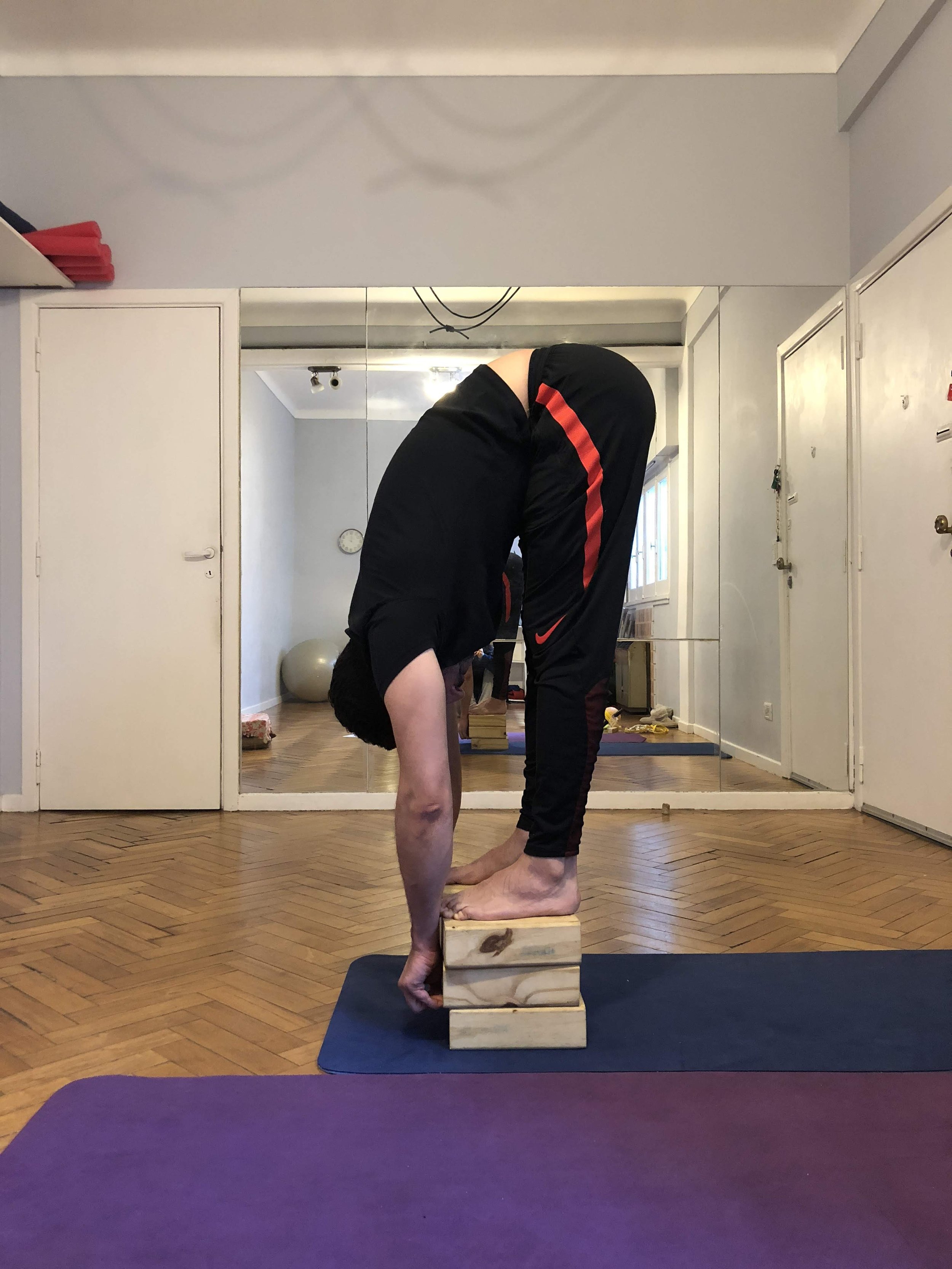 Person performing a forward fold on wooden blocks, bending forward with hands touching the floor in a well-lit room with wooden floor and a mirror background.