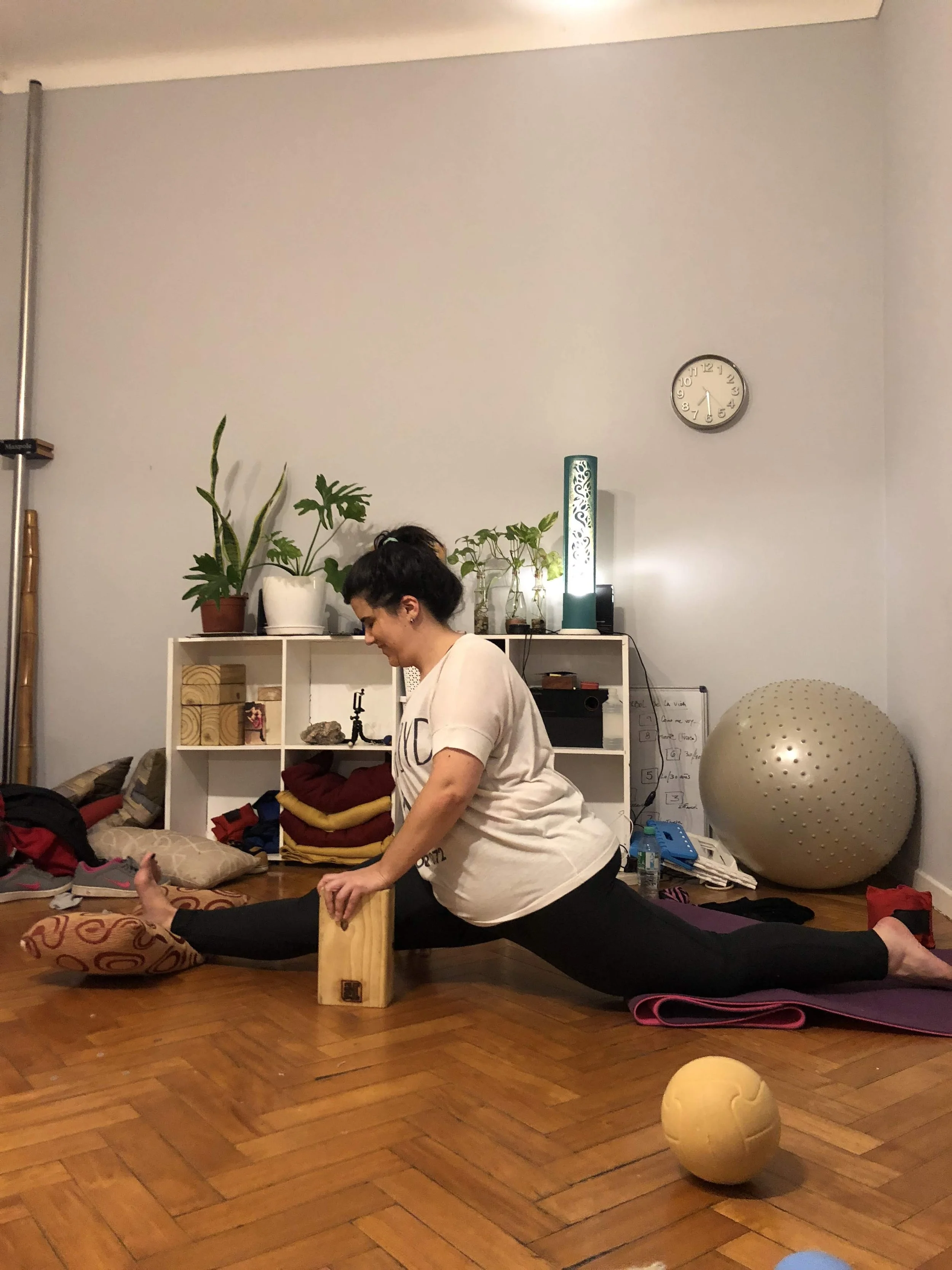 A woman performing a split stretch on a yoga mat in a room with wooden flooring, using a wooden block for support, surrounded by exercise equipment and houseplants.