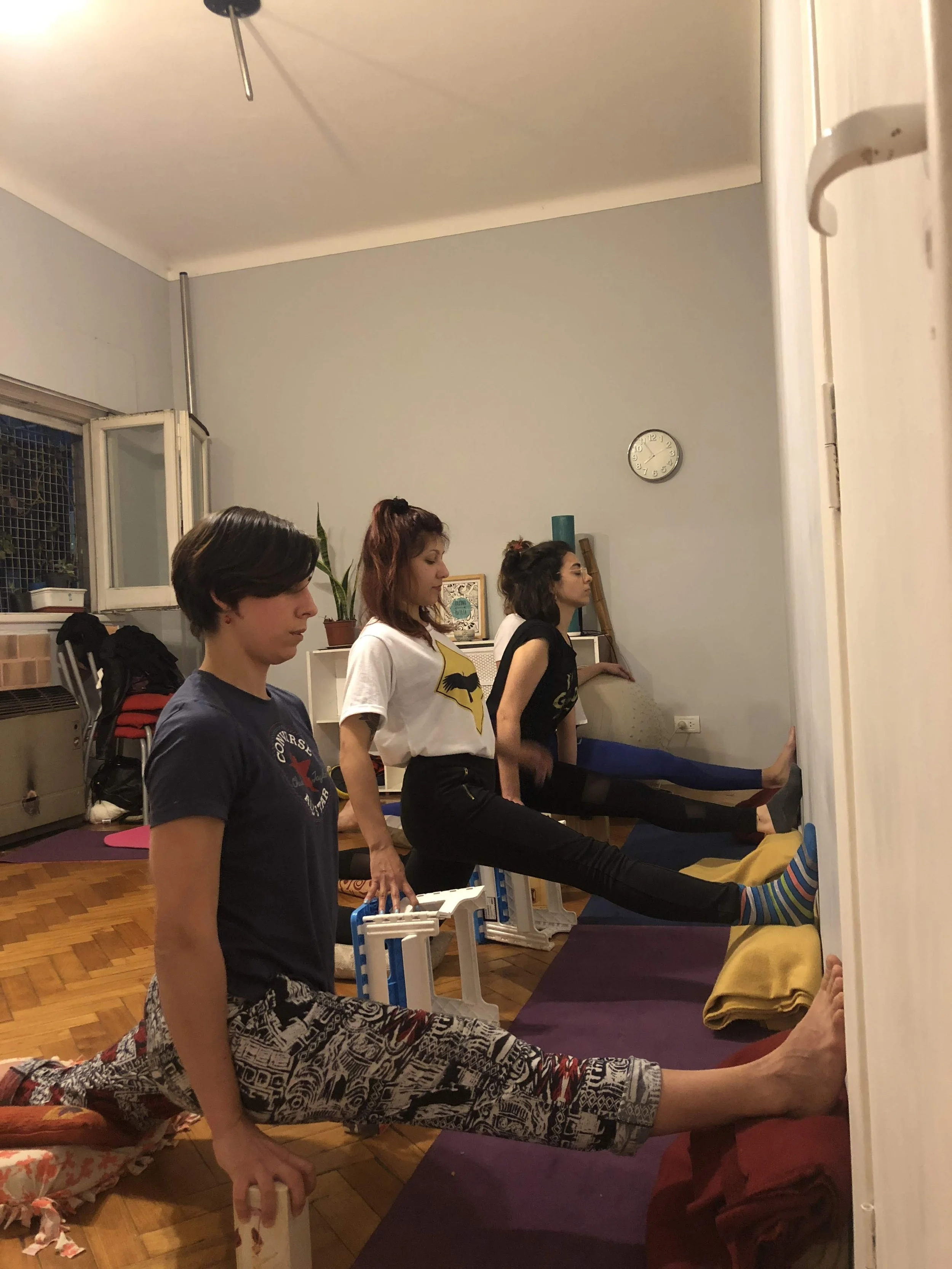 Three women stretching their splits in a living room, with one woman in the foreground holding a foam roller, and two women at the wall doing leg stretches.