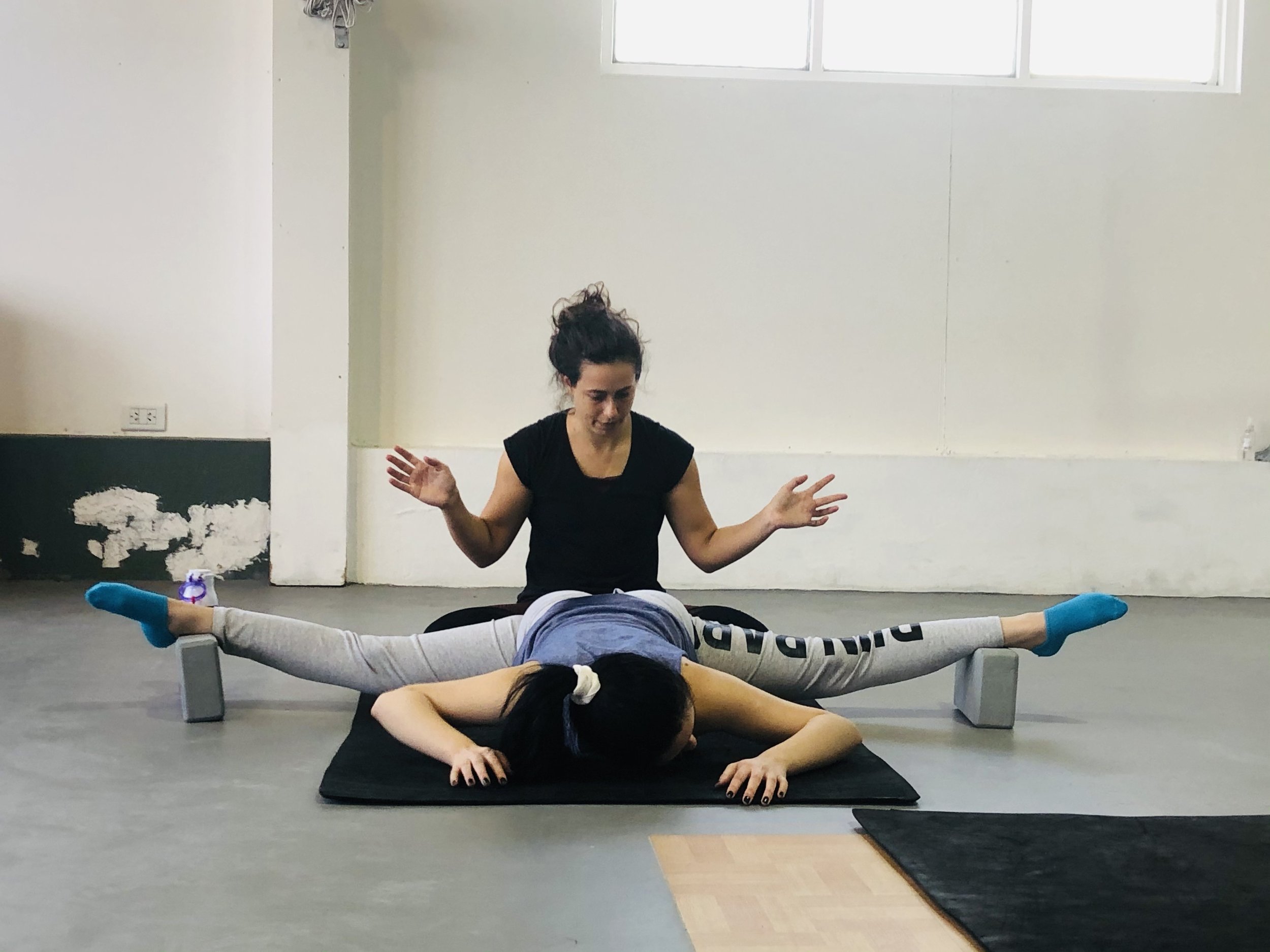 A woman is lying face down on a black mat with legs spread performing a leg stretch and feet resting on yoga blocks, while a instructor kneels behind her providing instructions for deepening the pancake stretch or middle split