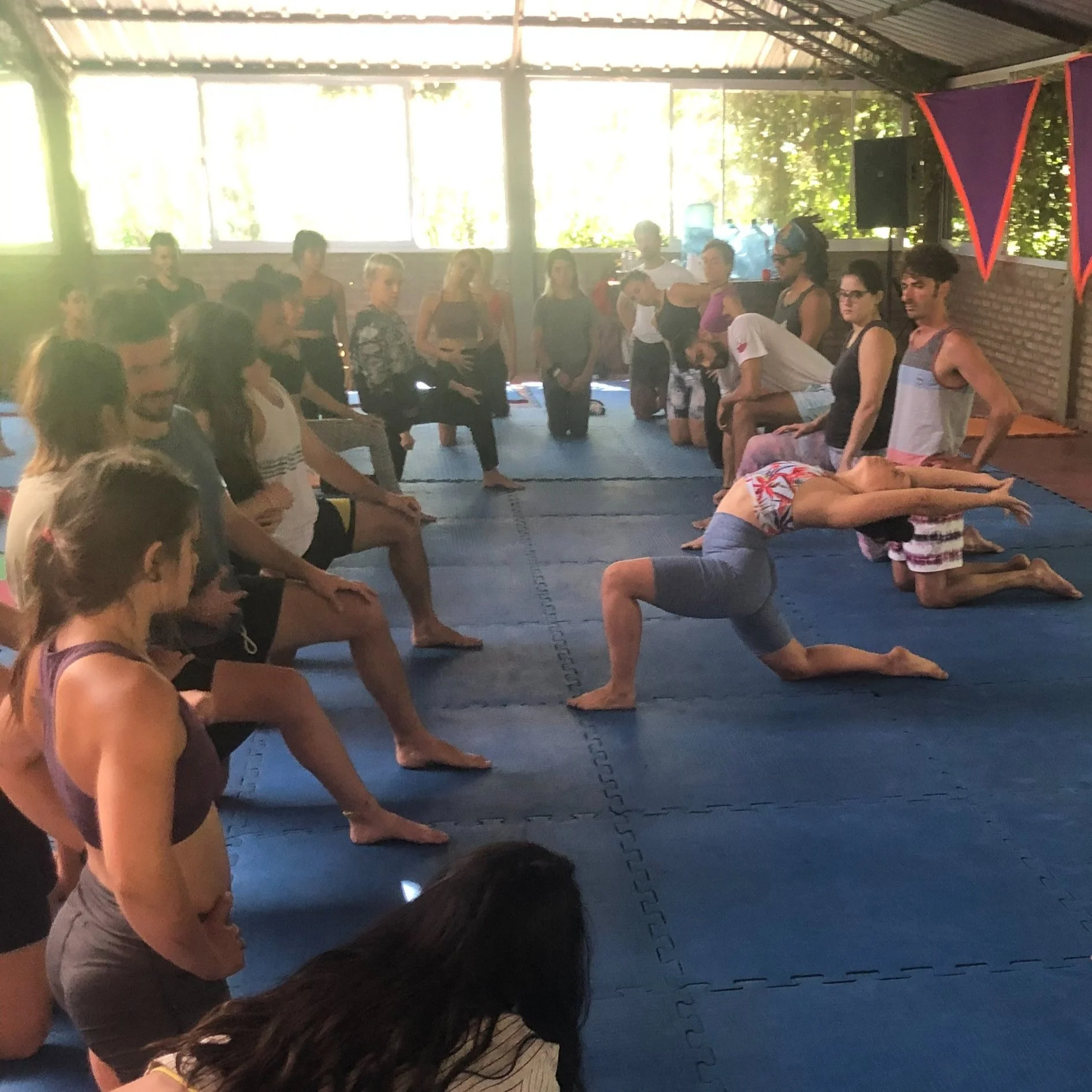 A group of people in a yoga class, with some practicing yoga poses and others observing, inside a bright indoor studio with blue mats and red and purple banners.