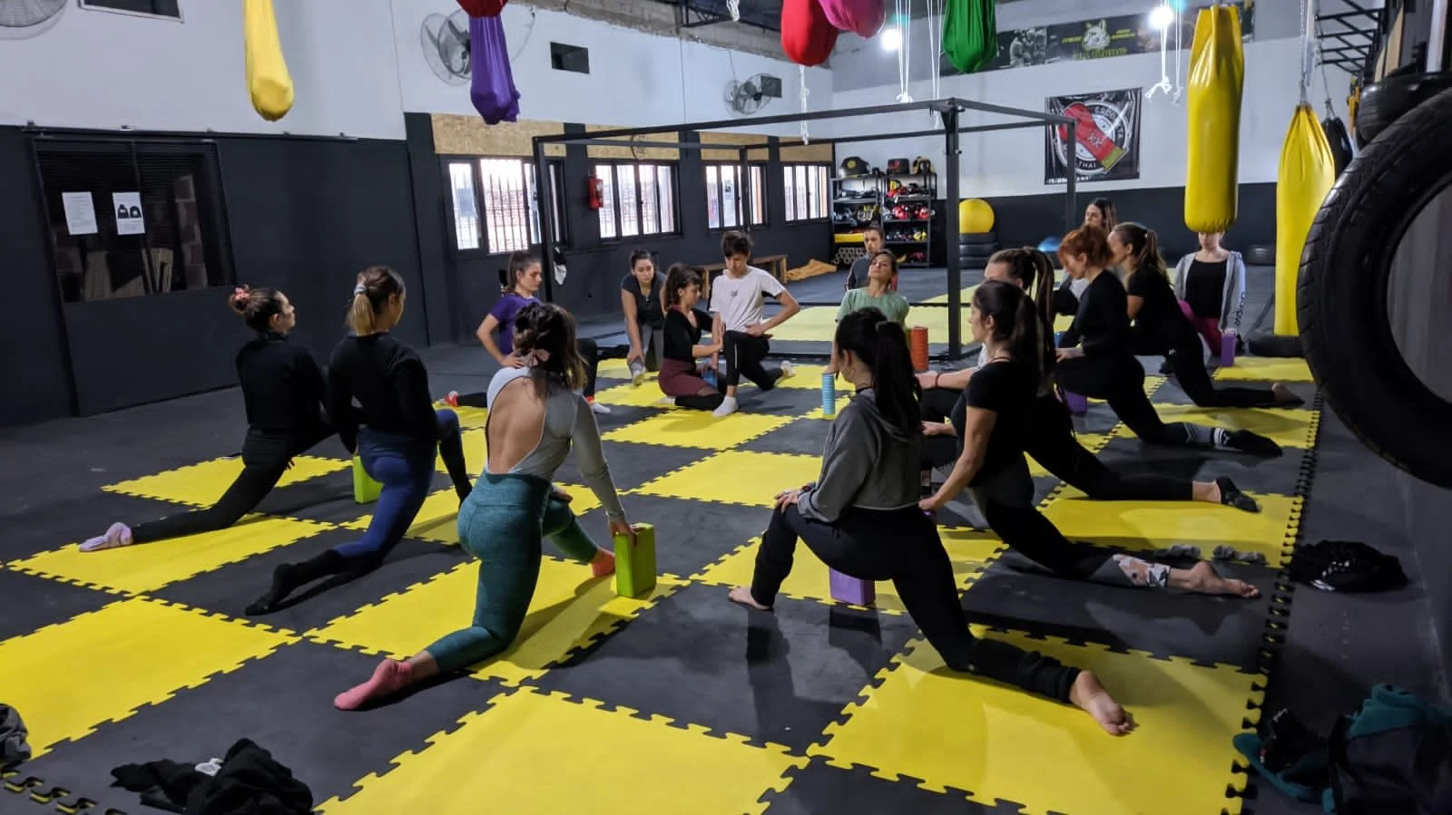 Group of women participating in a yoga or stretching class in a gym with yellow and black mats, hanging punching bags, and fitness equipment.