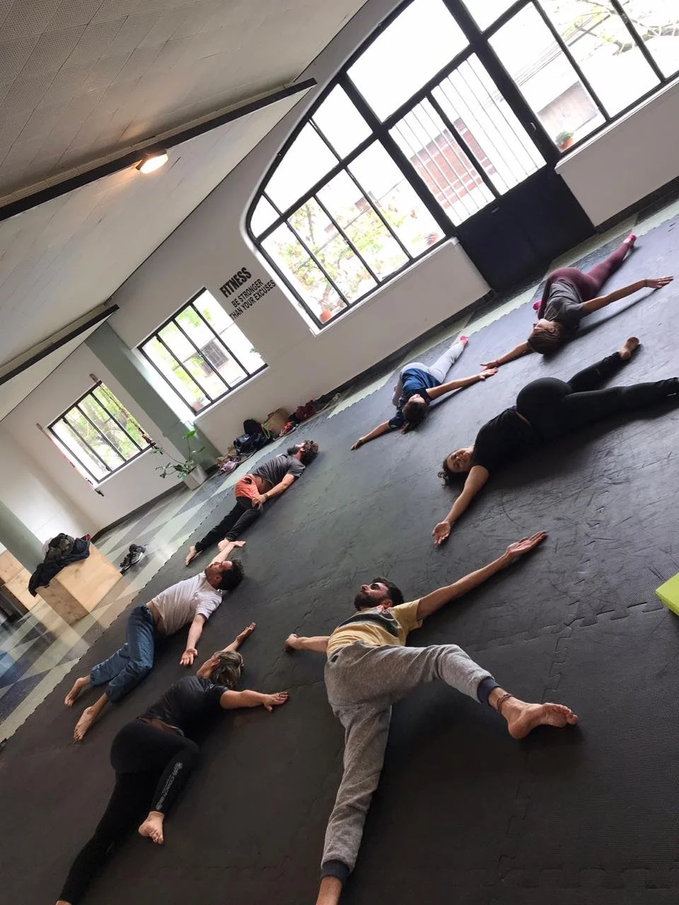 Group of people lying on a black mat in a spacious workout studio with large windows, practicing yoga or back stretching exercises.