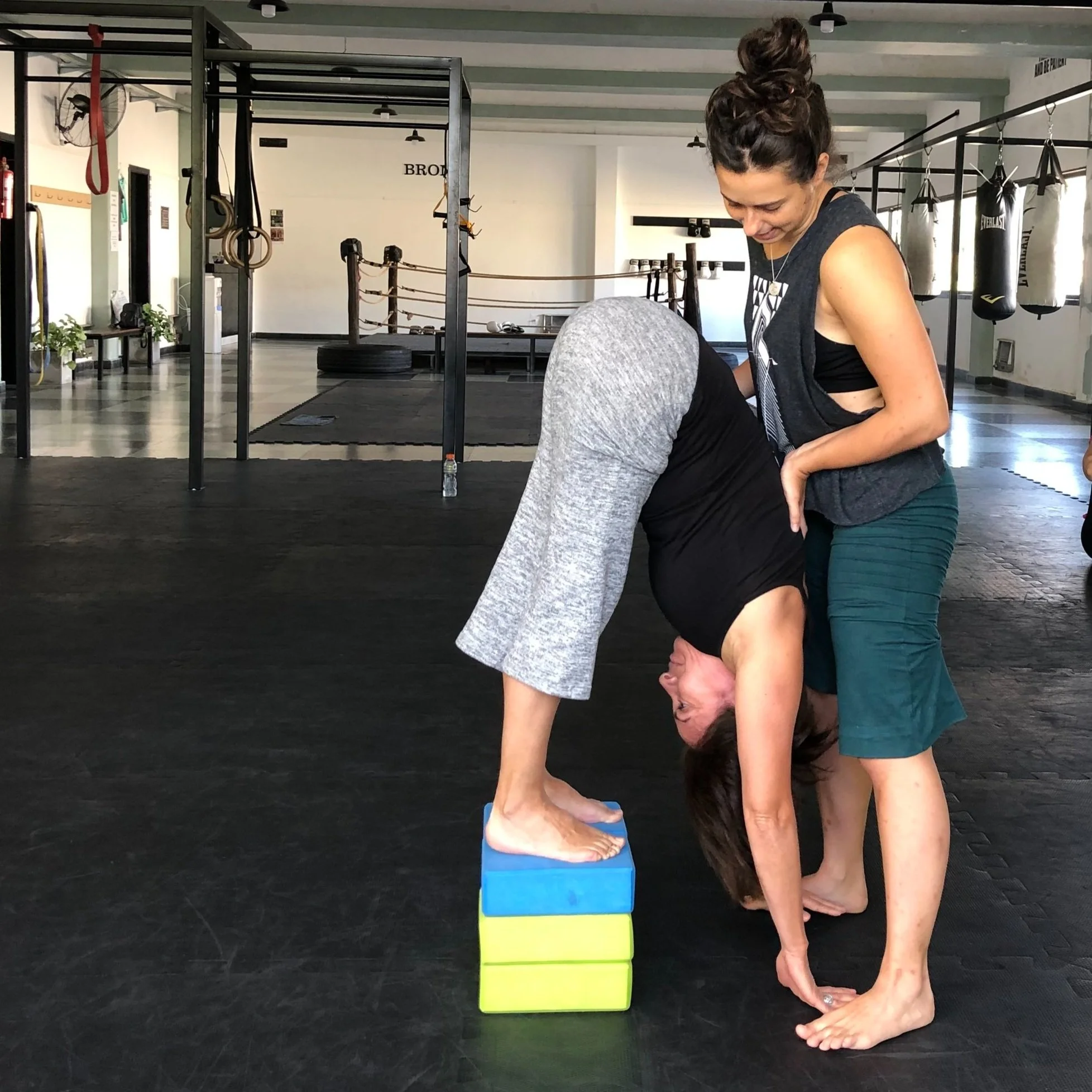 Two women practicing forward bends in a gym, one woman is upside down in a forward bend with her head touching the floor on top of foam blocks, and the other woman is standing and supporting her.