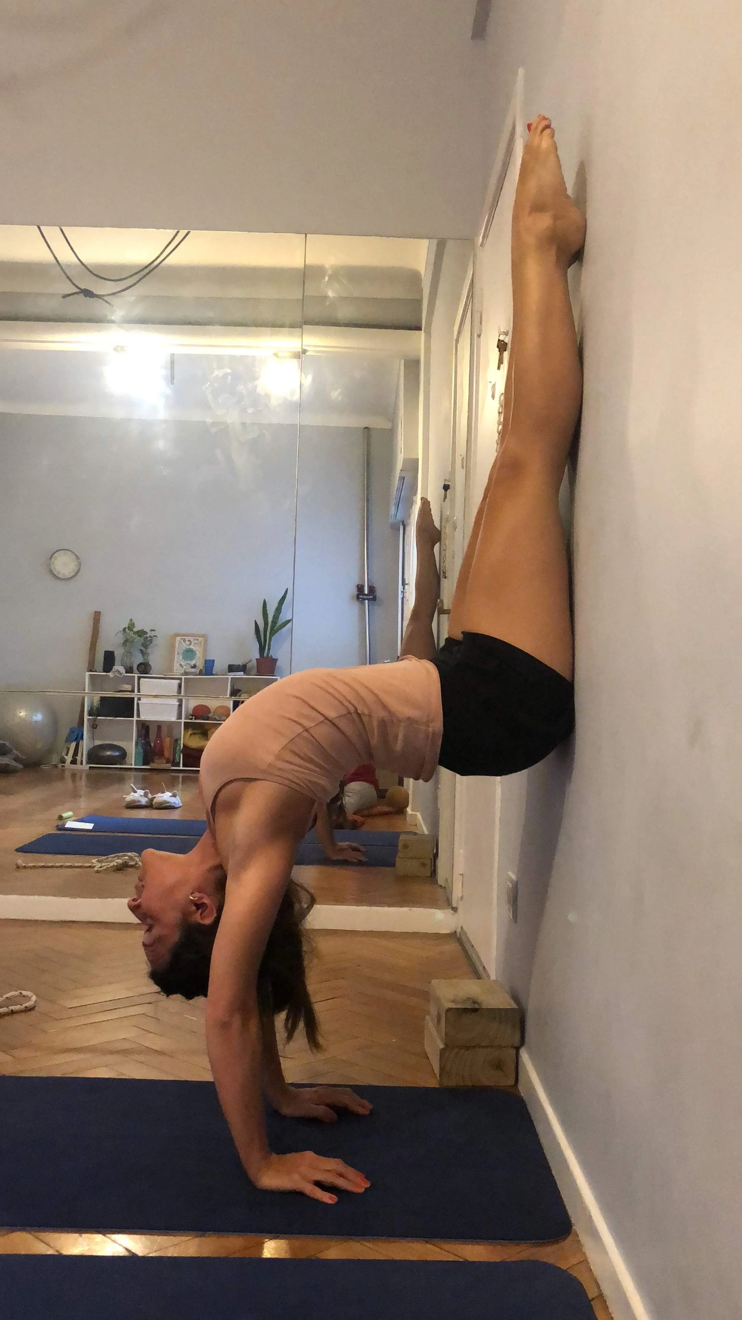 A woman practicing handstands with backbend in a yoga pose against a wall in a room with wooden floor and minimalist decor.
