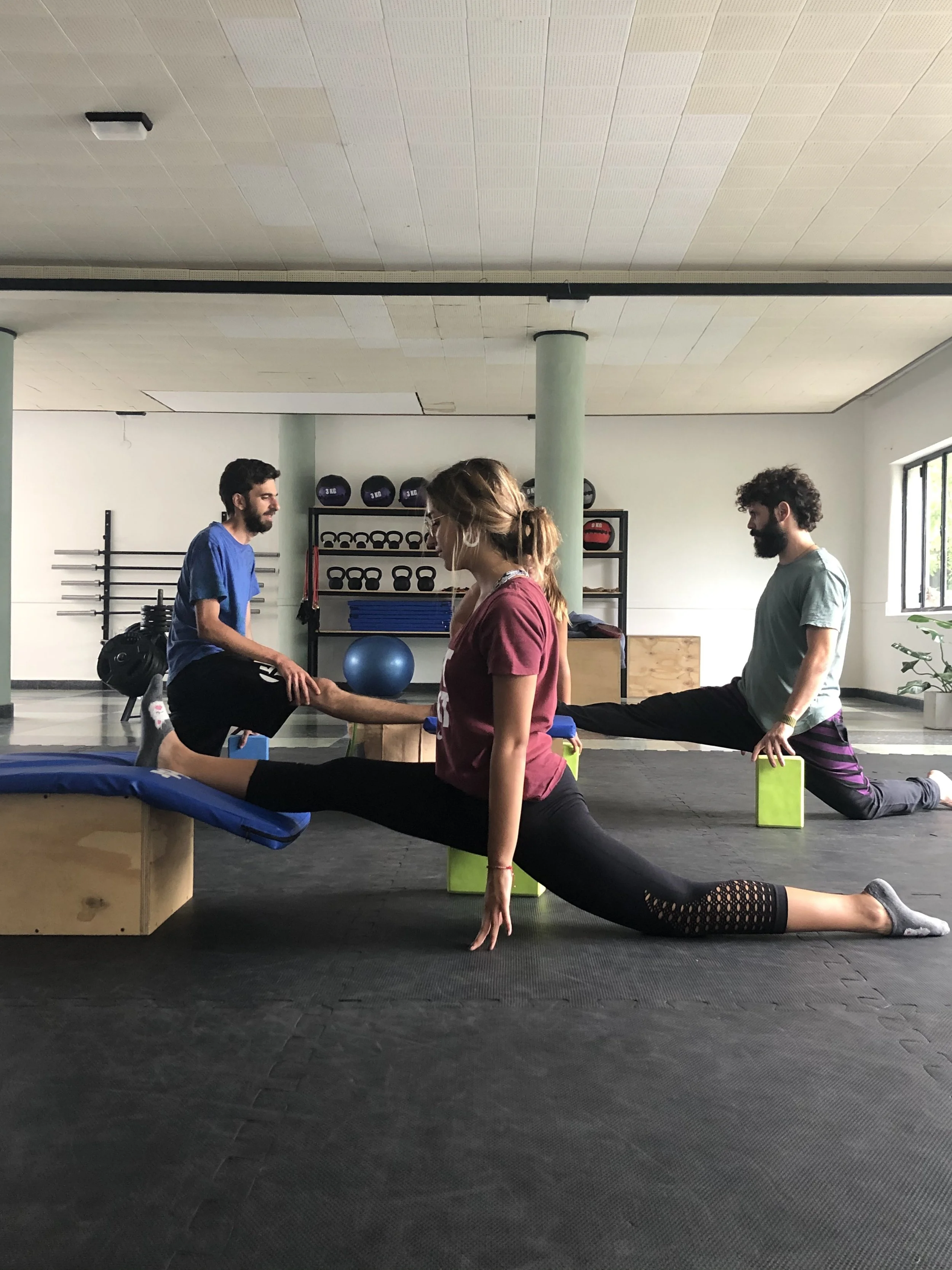 Three people in a gym practicing stretches or yoga. Two men are on one knee, each with one leg extended on a bench or block, while a woman in the center is performing a split stretch forward with her hand touching the person on the left. Gym equipment such as medicine balls, kettlebells, and exercise mats are visible in the background.