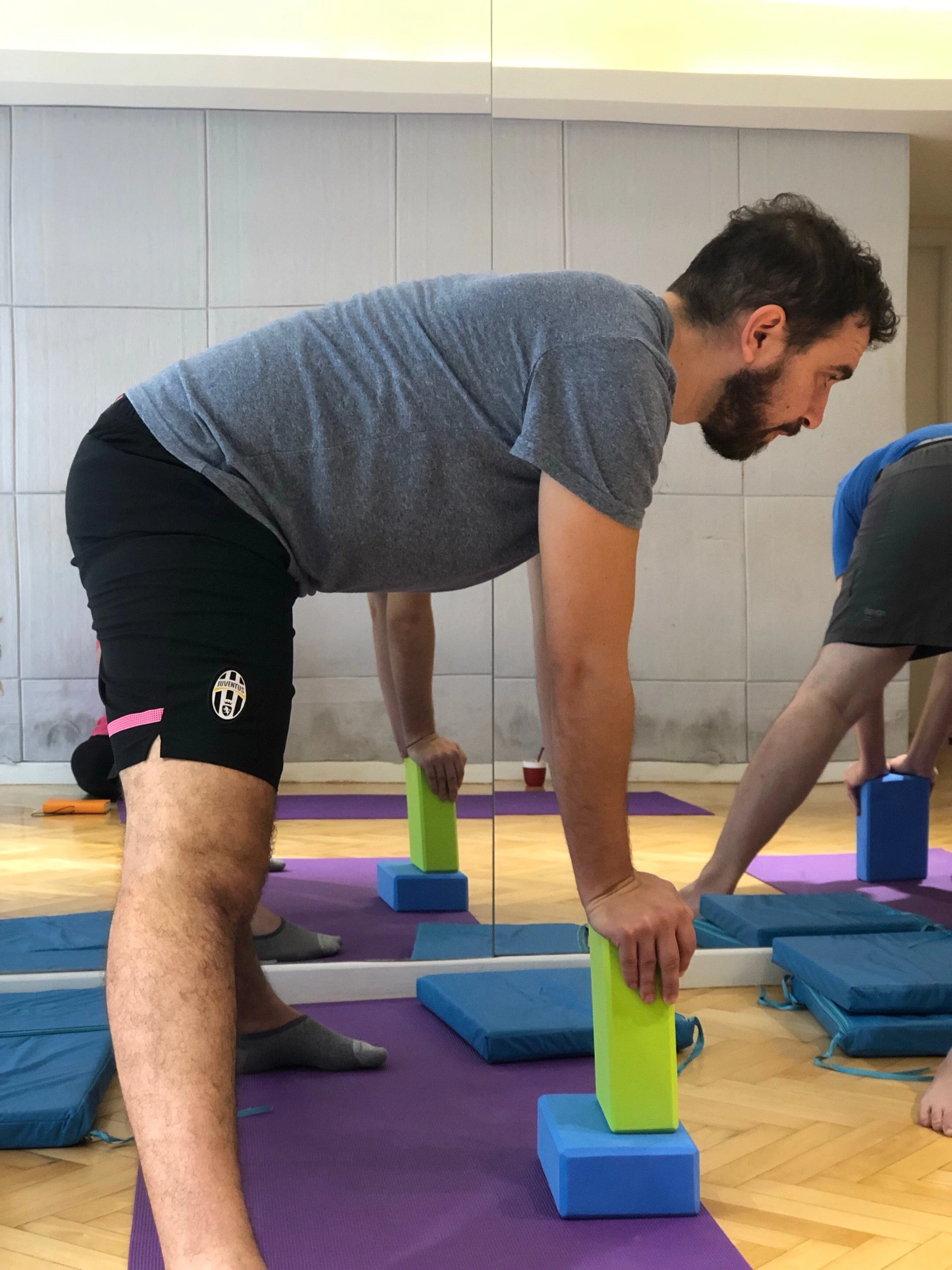 Man doing yoga on a purple mat, with blocks and other participants practicing in the background. It looks like he is releasing the hips to go into a middle split. 