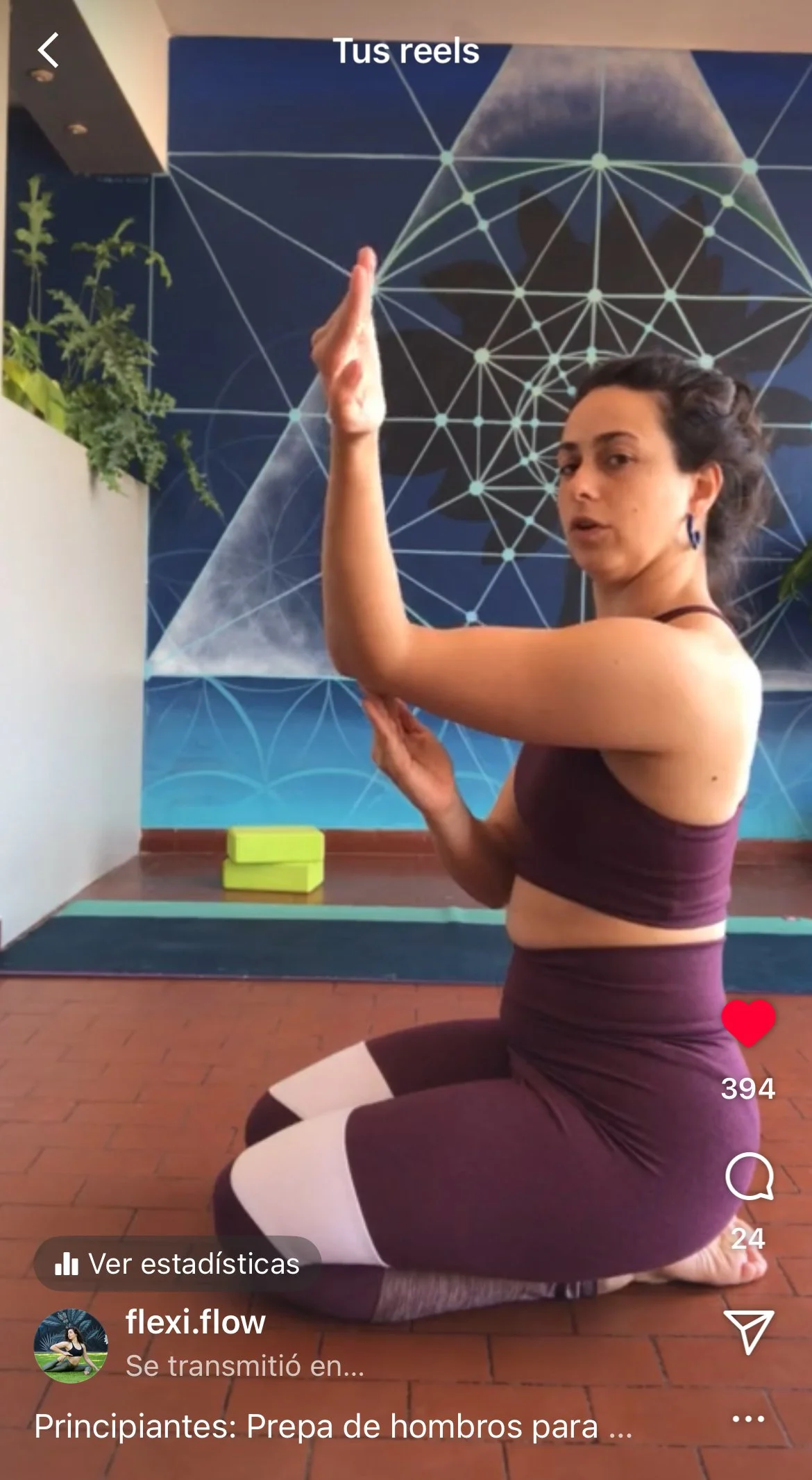 A woman indoors teaching an active flexibility class online via zoom, kneeling on a yoga mat with her right arm raised and bent at the elbow, her hand near her head. She is wearing a dark sports bra and matching leggings with white accents.