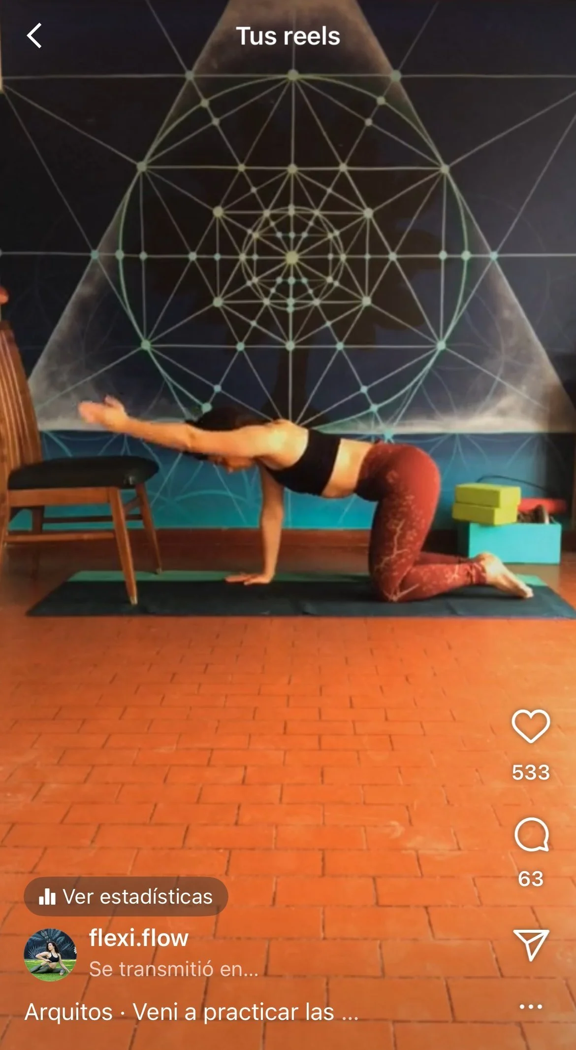 A woman instructing flexibility or stretching lessons to students via instagram live, performing a modified plank pose with one arm resting on a chair and her arm extended forward, against a wall with geometric patterns.