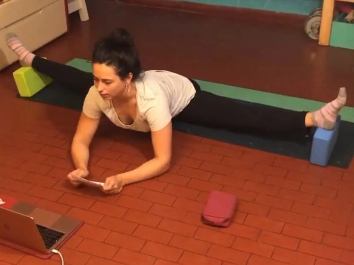 Woman practicing yoga in middle splits position with her legs supported by foam blocks while looking at a laptop on the floor. She is instructing students on how to get their middle splits