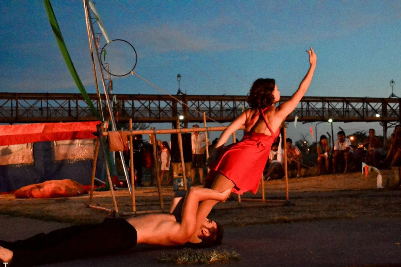 A woman in a red dress is balanced on a man's feet in an outdoor setting during dusk, with a crowd and a wooden bridge in the background.