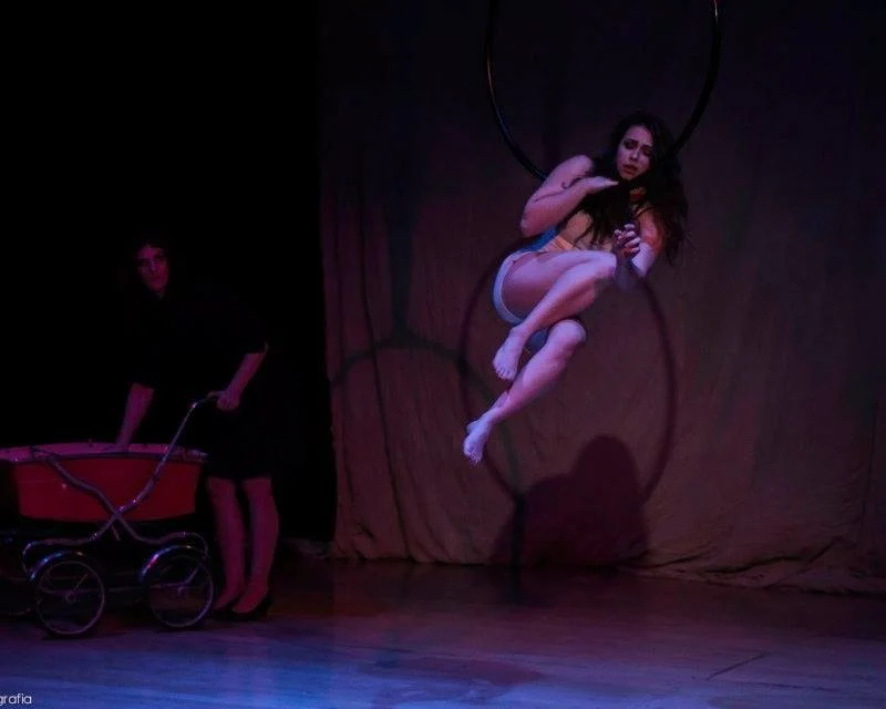 A woman performing an aerial act on a hoop, suspended in the air, with a man standing and holding a red vintage stroller in a dimly lit setting.