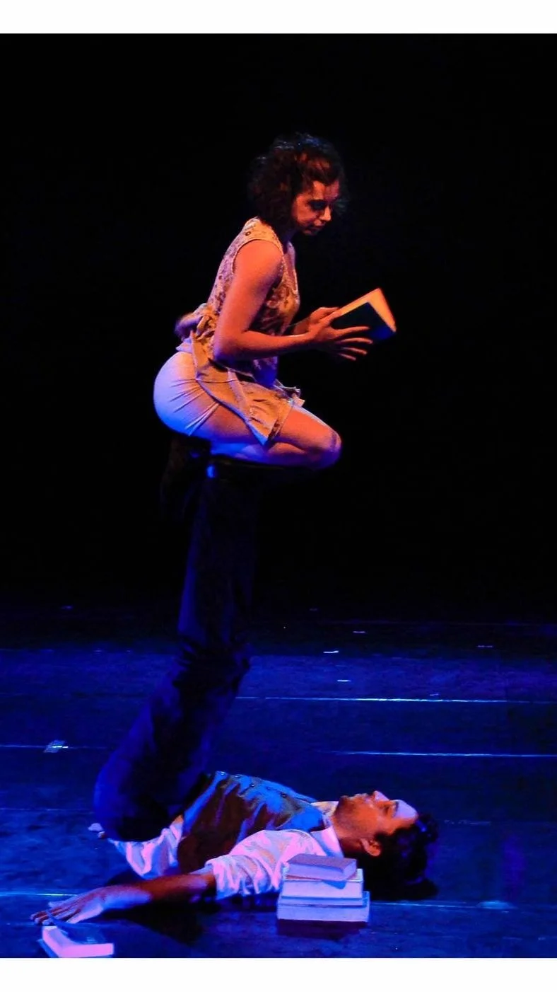 A woman standing on a man's legs who is lying on the ground; they are on a stage with books and a phone around them, and the background is dark with stage lighting.