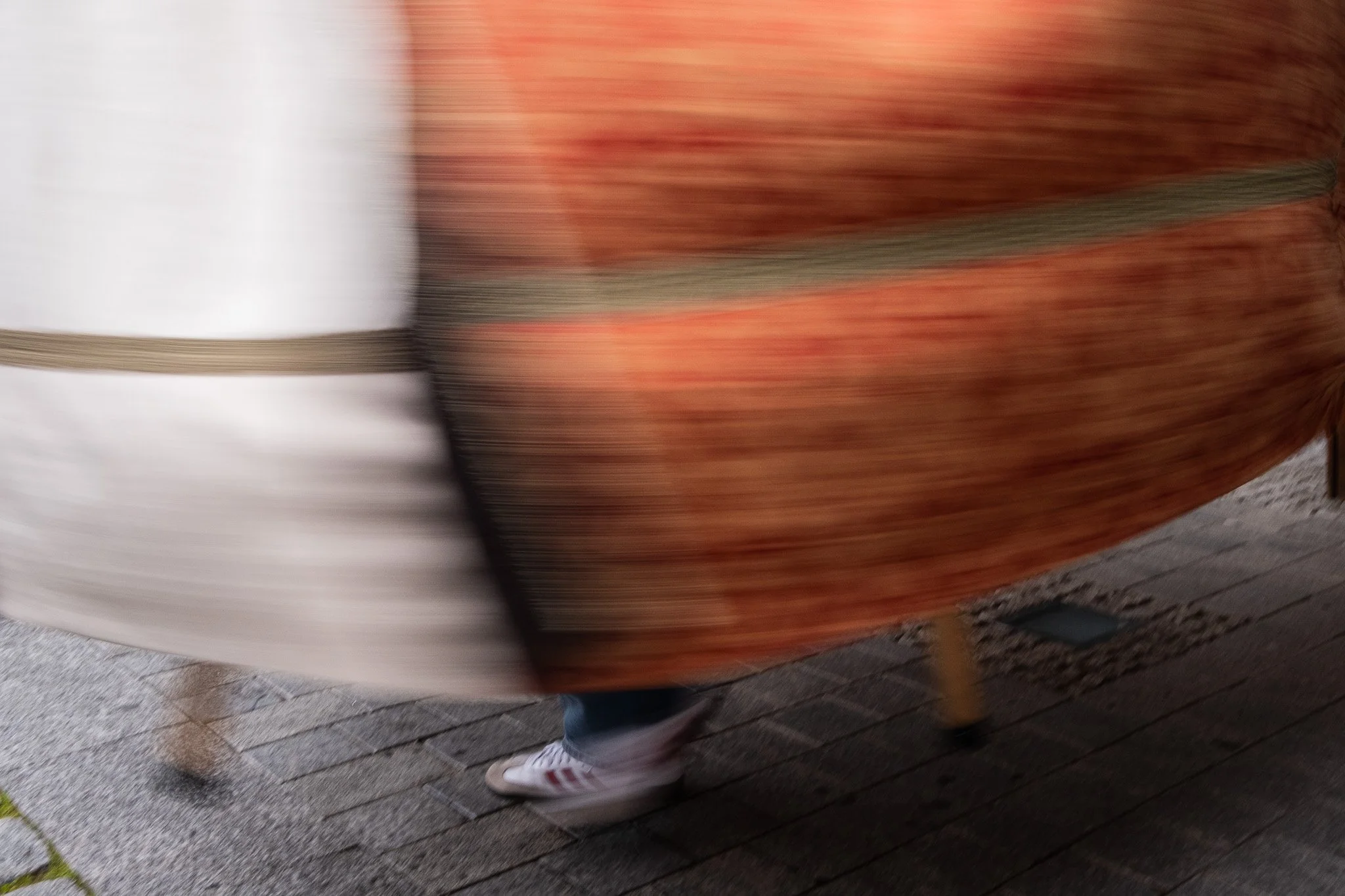 Blurred image of a person walking, wearing white Adidas sneakers and a flowing, colorful, patterned garment.