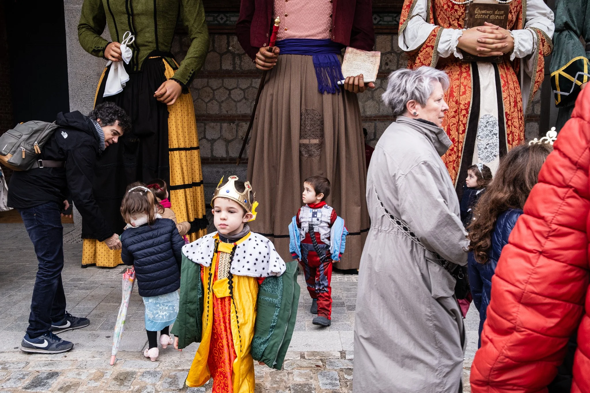 Children and adults gathered outside a building, some dressed in costumes, including a child wearing a king's costume with a crown and a regal cape, and other children dressed warmly in jackets. Adults are engaged with the children, with one woman in