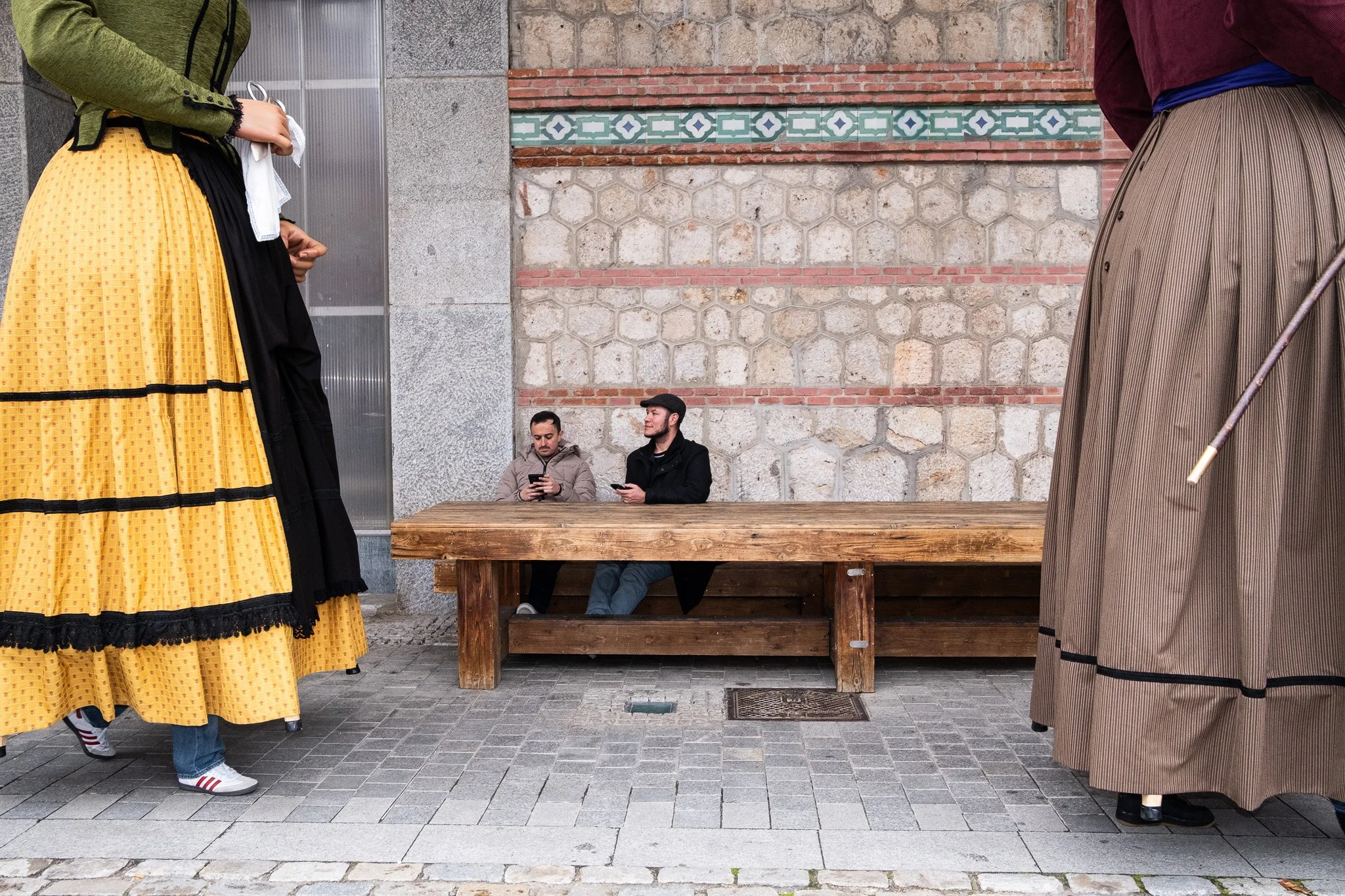 Two men sitting at a wooden bench looking at their phones, surrounded by two women in traditional dresses standing nearby, against a stone and brick wall background.