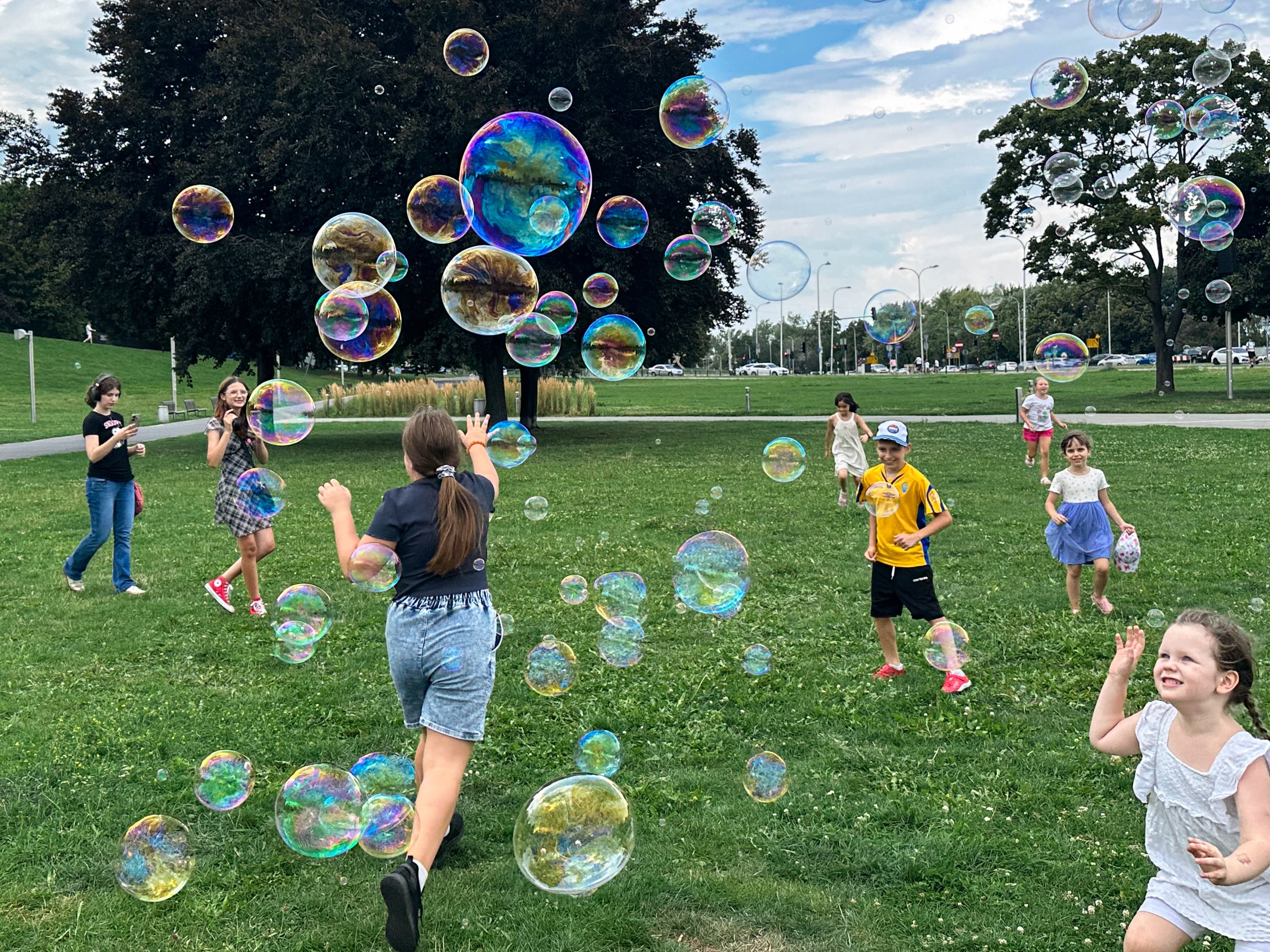 Children playing with soap bubbles in a park on a cloudy day, with trees and a parking lot in the background.