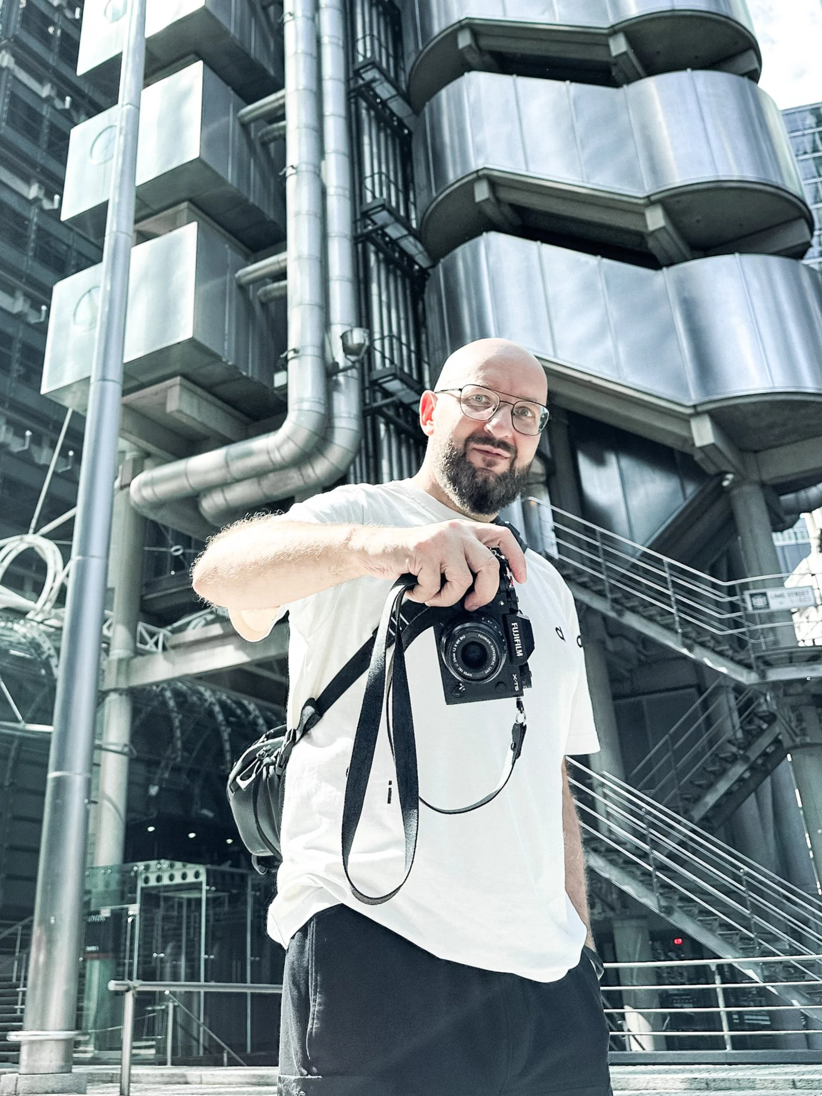 A man with glasses, a beard, and a bald head holding a camera is standing outdoors in front of a modern metal building with pipes and balconies.
