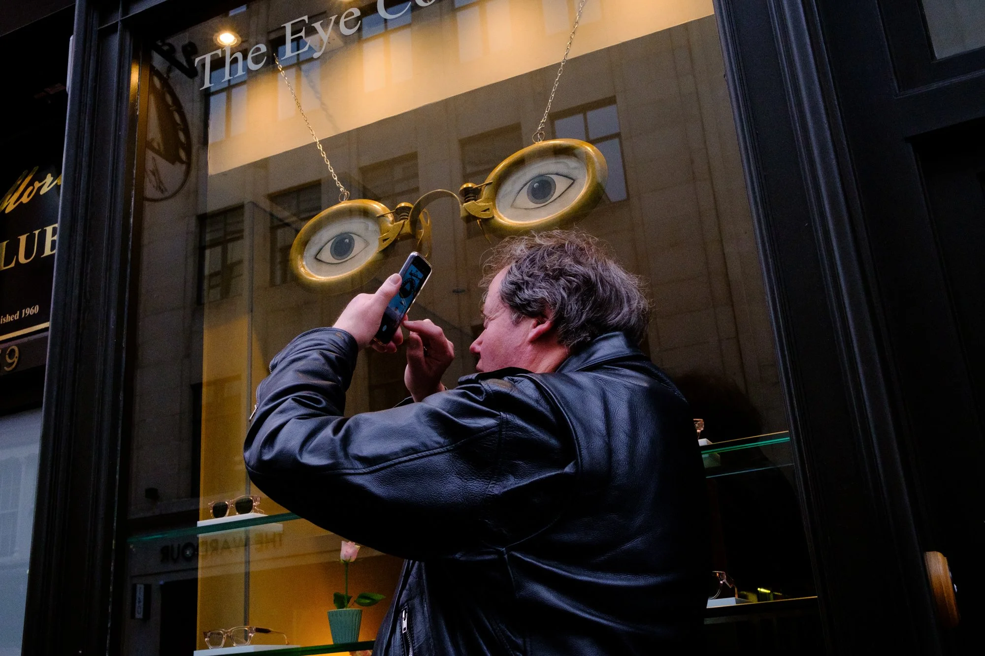 A man in a black leather jacket taking a photo of a storefront window display featuring a pair of glasses with eyes and a chain, with the store name "The Eye" partially visible.