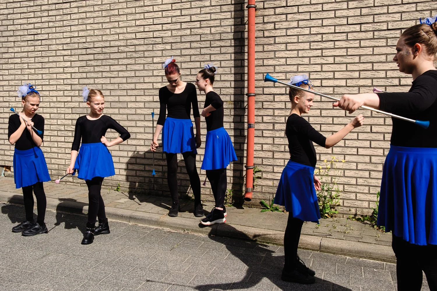 Young girls in black tops and blue skirts outside, holding batons, practicing a routine or dance with some standing and some interacting near a brick wall.