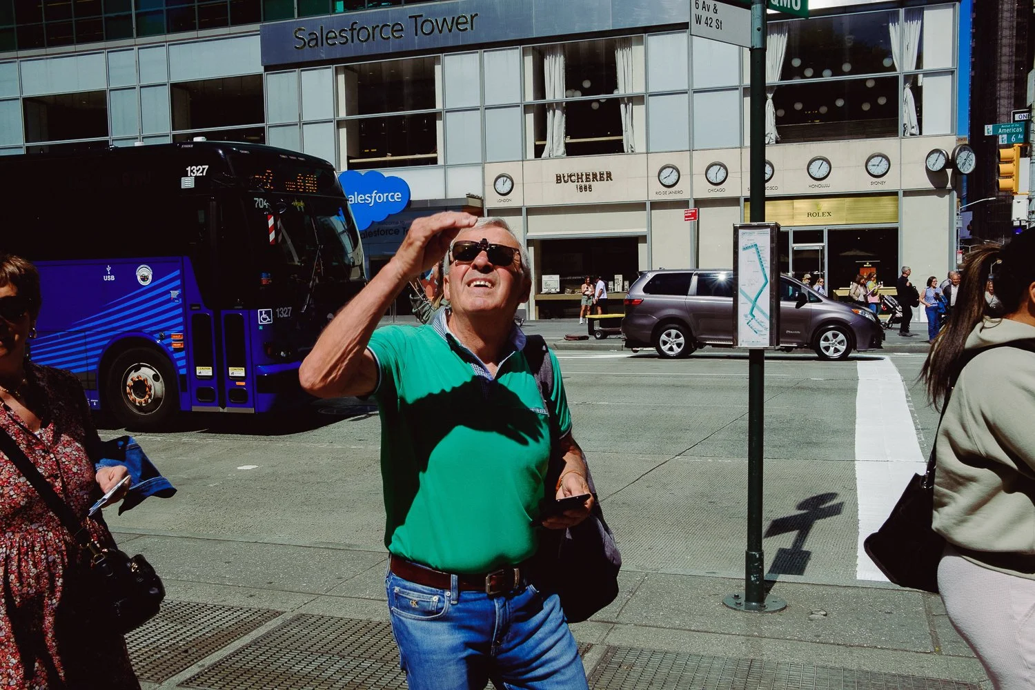 A man with sunglasses wearing a green shirt, standing on a city sidewalk, shielding his eyes from the sun, with a Salesforce Tower building in the background.