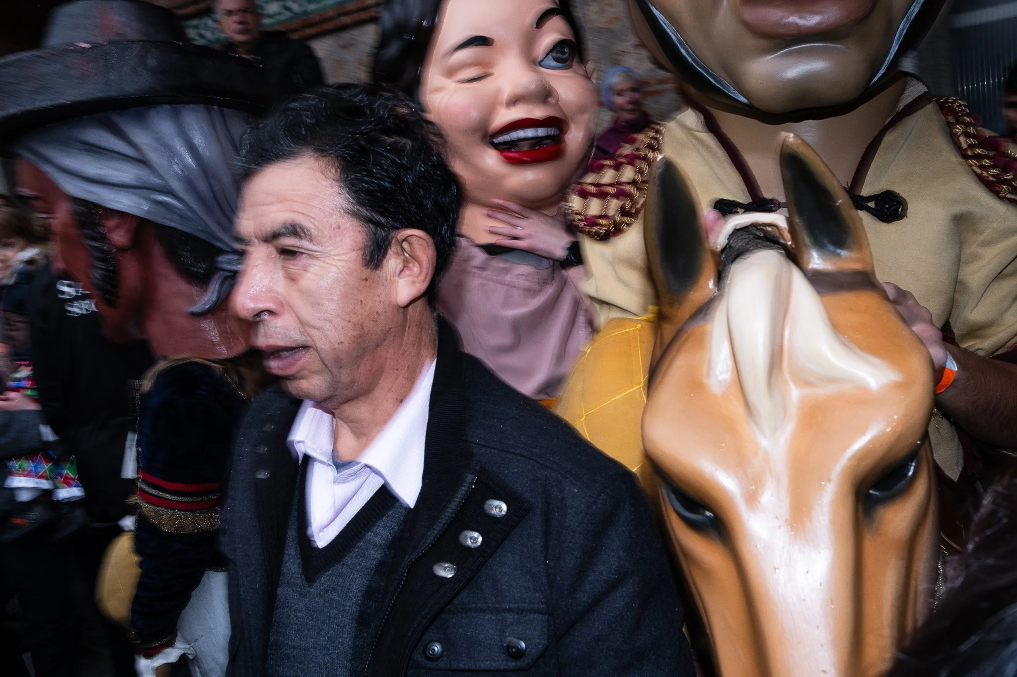 People in a crowded setting with two individuals wearing large masks; one mask depicts a caricatured face with exaggerated features and the other is a horse mask.