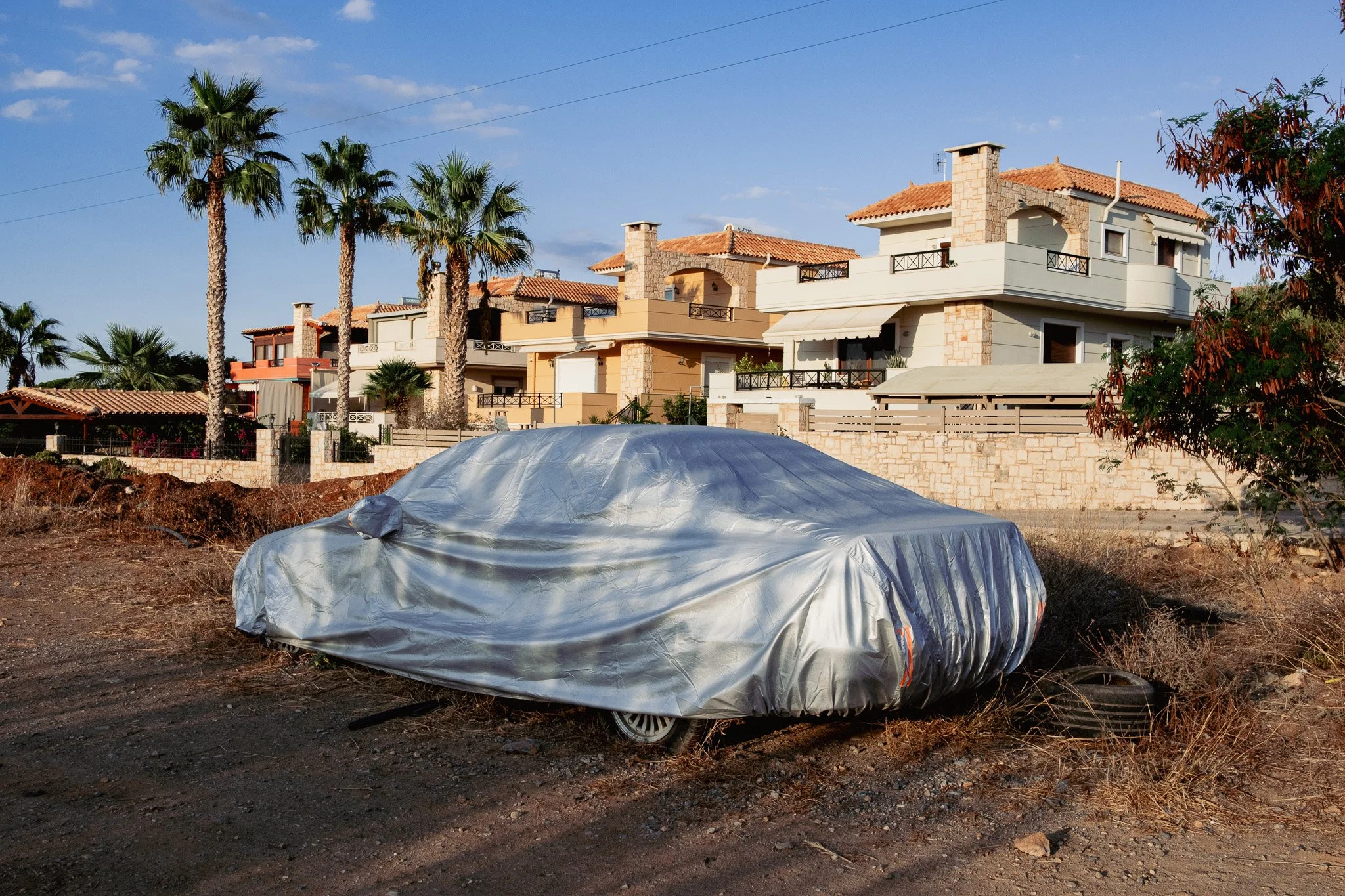 A car covered with a silver protective cover parked in front of modern multi-story houses with orange tile roofs and palm trees under a blue sky.