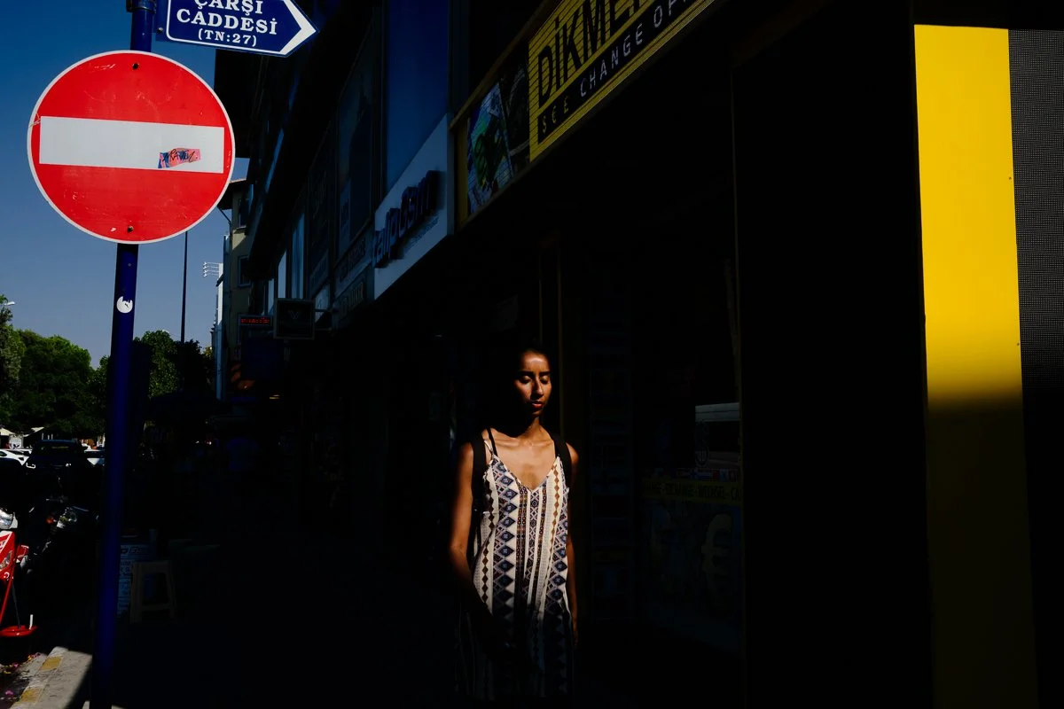 A young woman walking on a city street with street signs and storefronts, partly in shadow with bright sunlight on her face. 