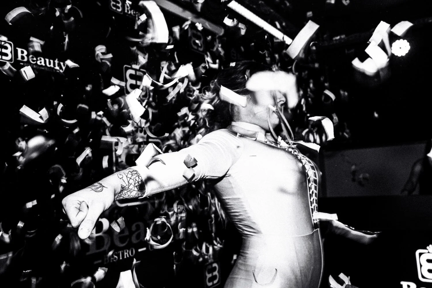 A person with tattoos on their arm and tattoos on their neck throwing their arm outward amidst falling confetti, with a background of a wall full of small paper items and text, all in black and white.