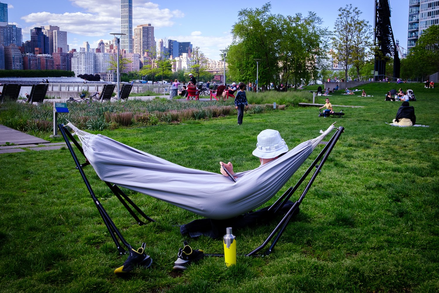 Person relaxing in a hammock on the grass in a city park with people sitting, walking, and playing in the background, and a city skyline with tall buildings behind them.