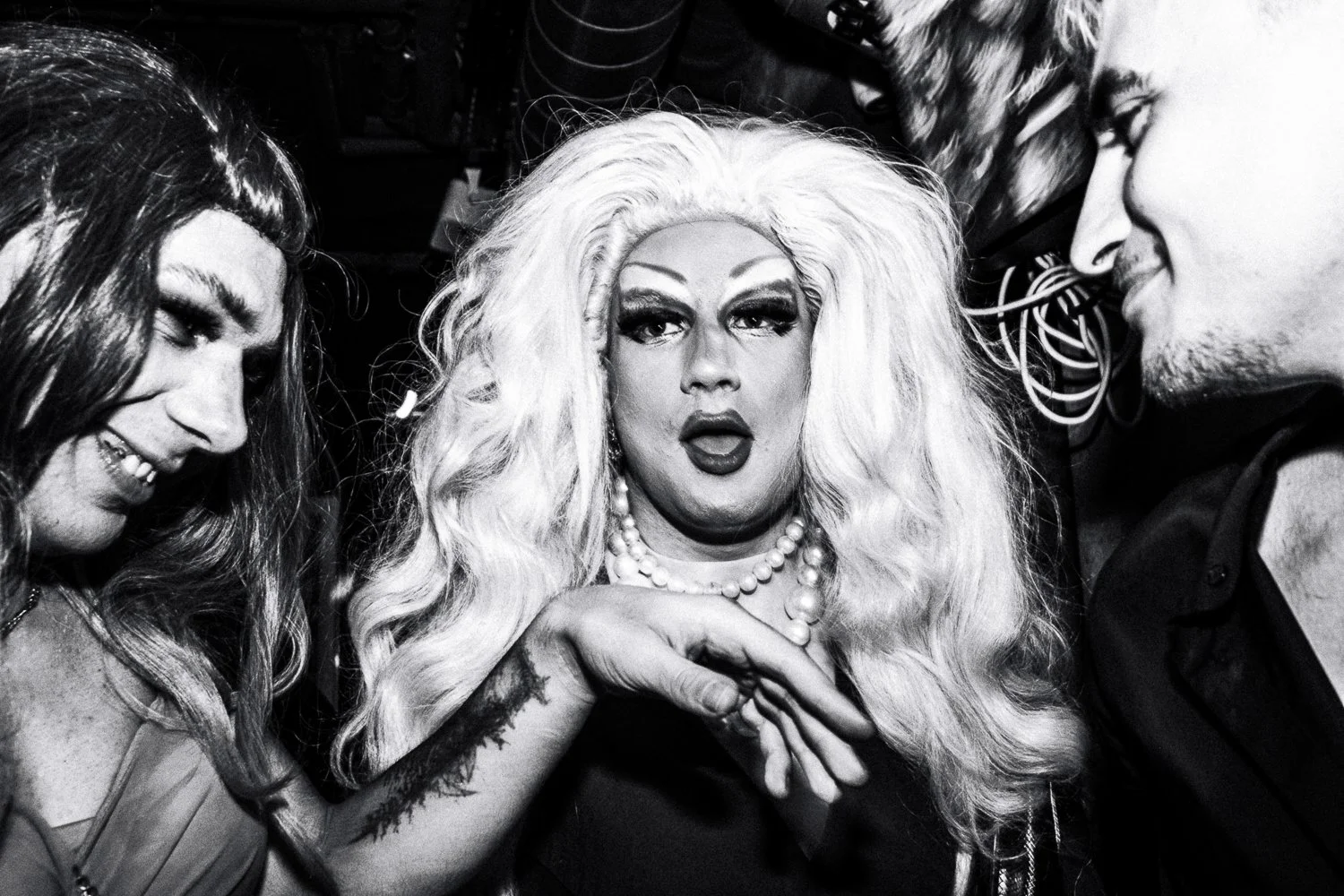 Three people, two men and a person with long blonde hair and large makeup, posing together in black and white photo.