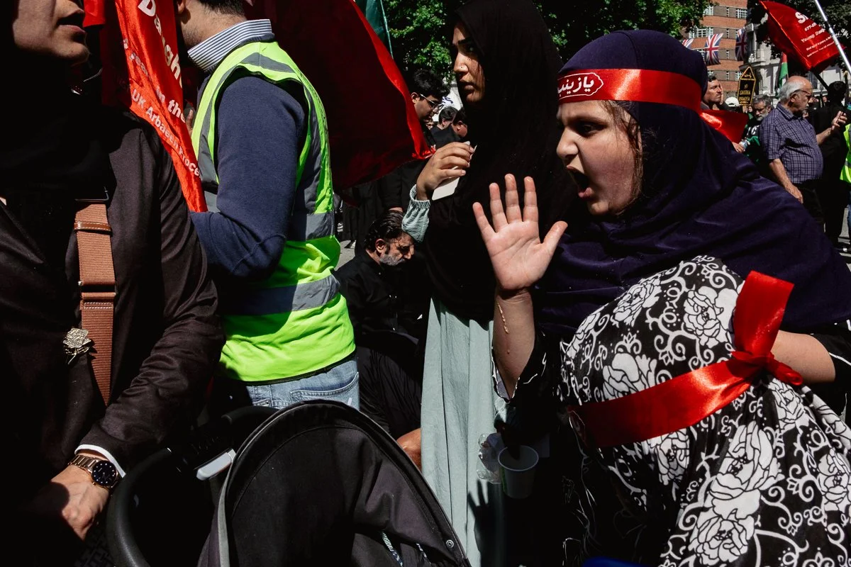 A woman wearing a red headband and black headscarf shouting at another person during a protest or demonstration, with many other protesters and flags in the background.