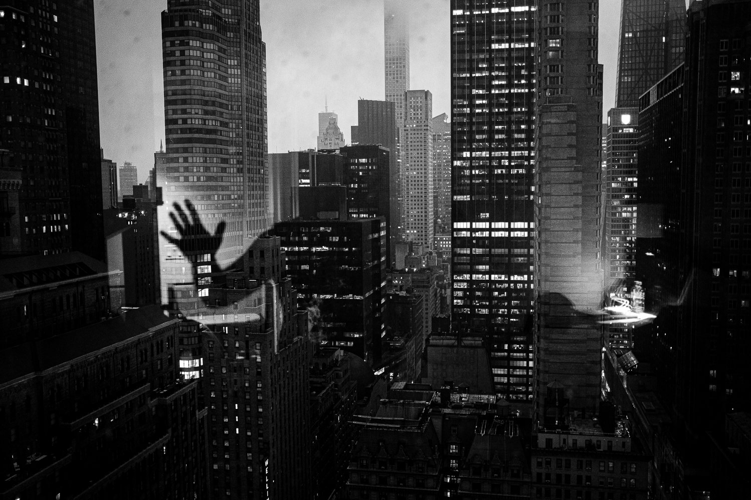 City skyline at night with numerous illuminated skyscrapers and a large window reflecting a person reaching out with an open hand.